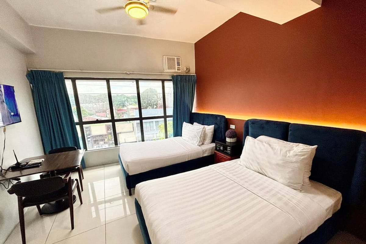 A hotel room with two beds, a desk with a chair, a TV, and a window with curtains.