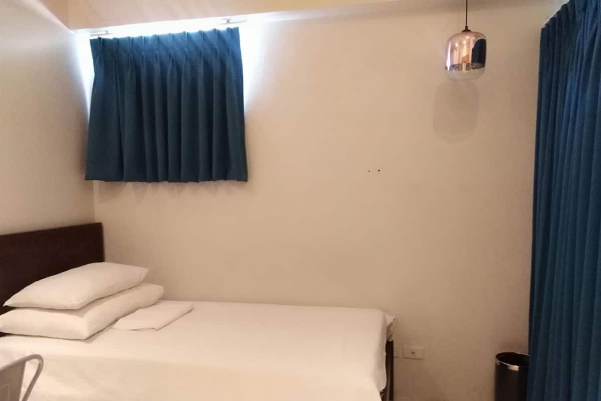A single bed with white linens, a dark headboard, a small trash bin, and a hanging light fixture.