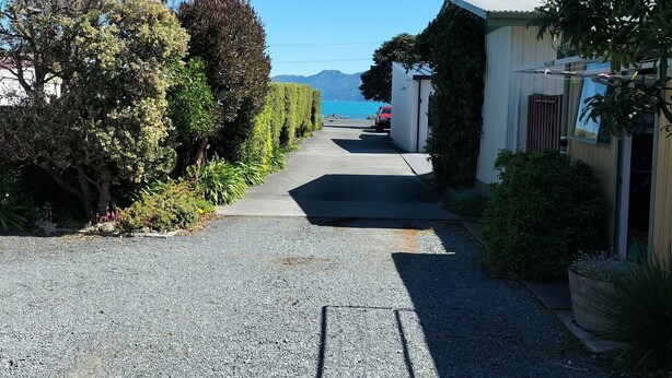 A residential street with a gravel driveway leading to a house with a red car parked in front. - Bay Cottages (Kaikoura)