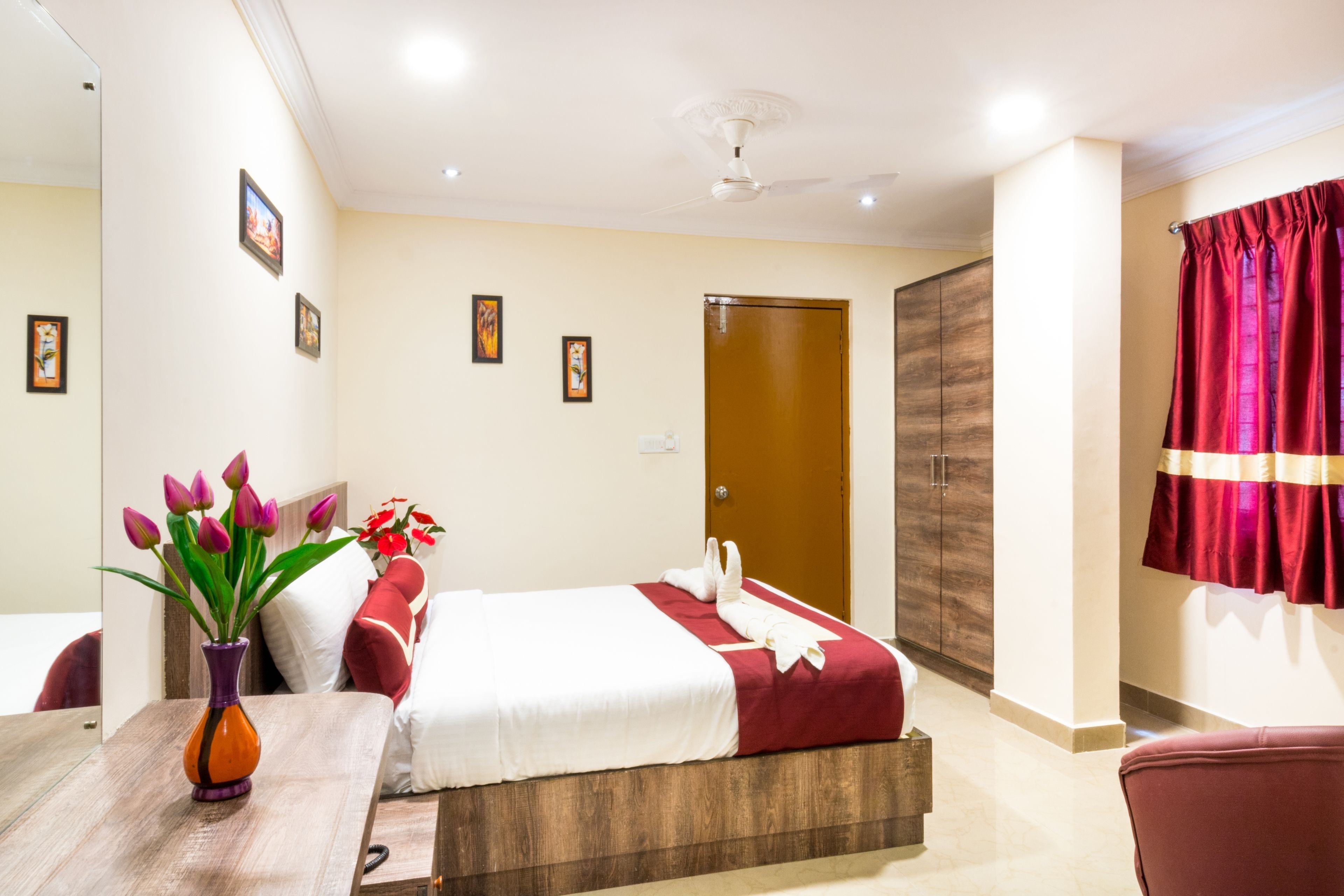 A hotel room with a bed, a wooden nightstand, a vase with flowers, and a red curtain.