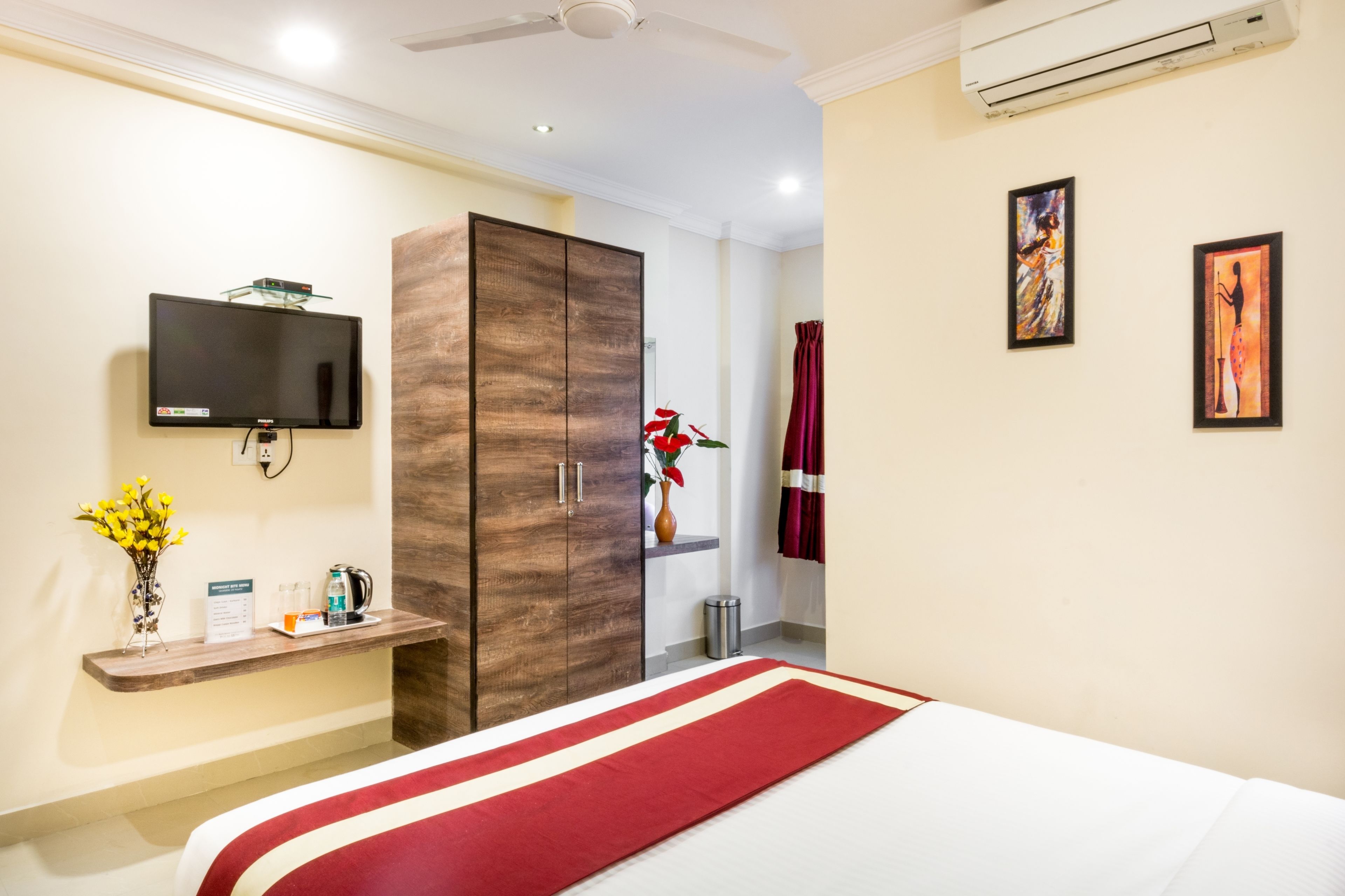 A hotel room with a bed, a TV mounted on the wall, a wardrobe, a small table with a vase of flowers, and a red and white striped area rug.