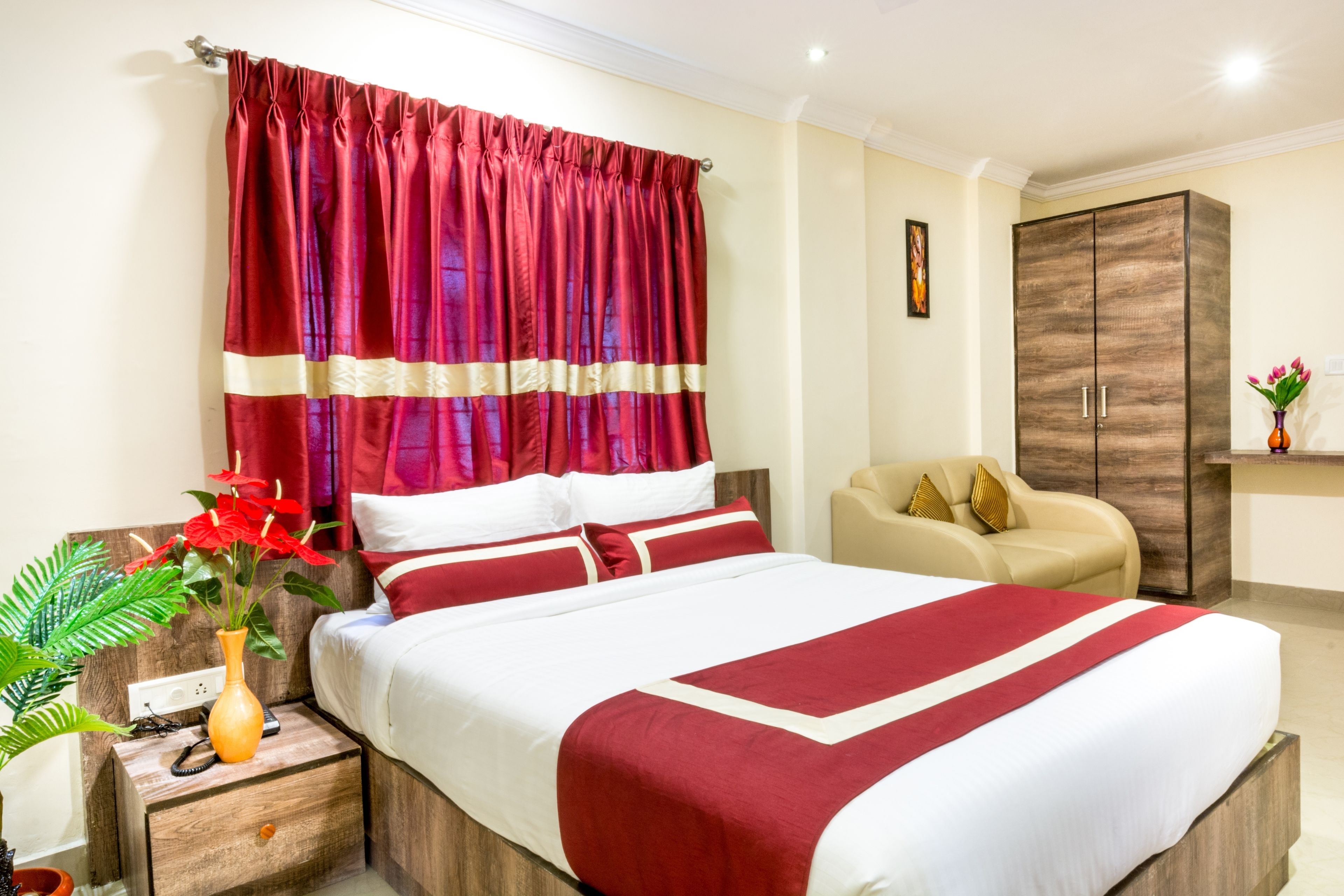 A neatly made bed with red and white bedding, a wooden nightstand with a vase of flowers, a beige armchair, and a wooden wardrobe.