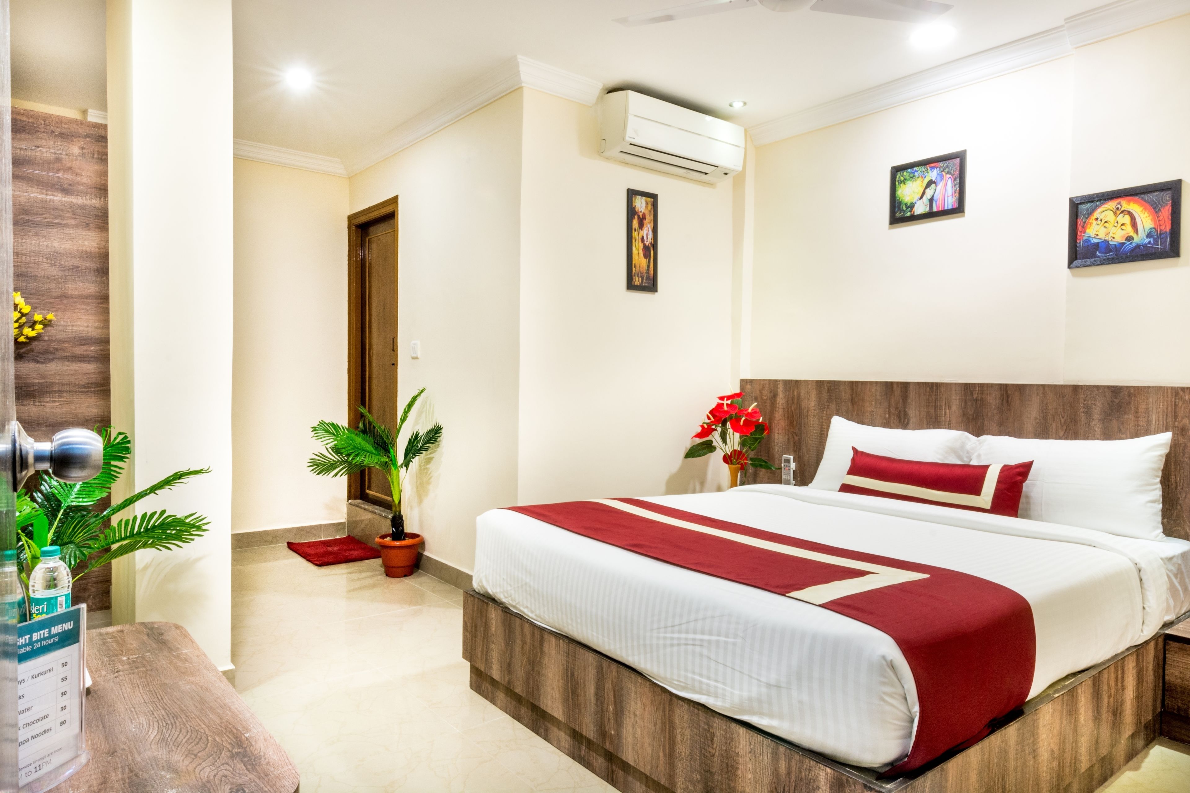 Habitación de hotel con cabecera de madera, una cama grande con ropa de cama blanca y roja, una planta en maceta y un aire acondicionado de pared.