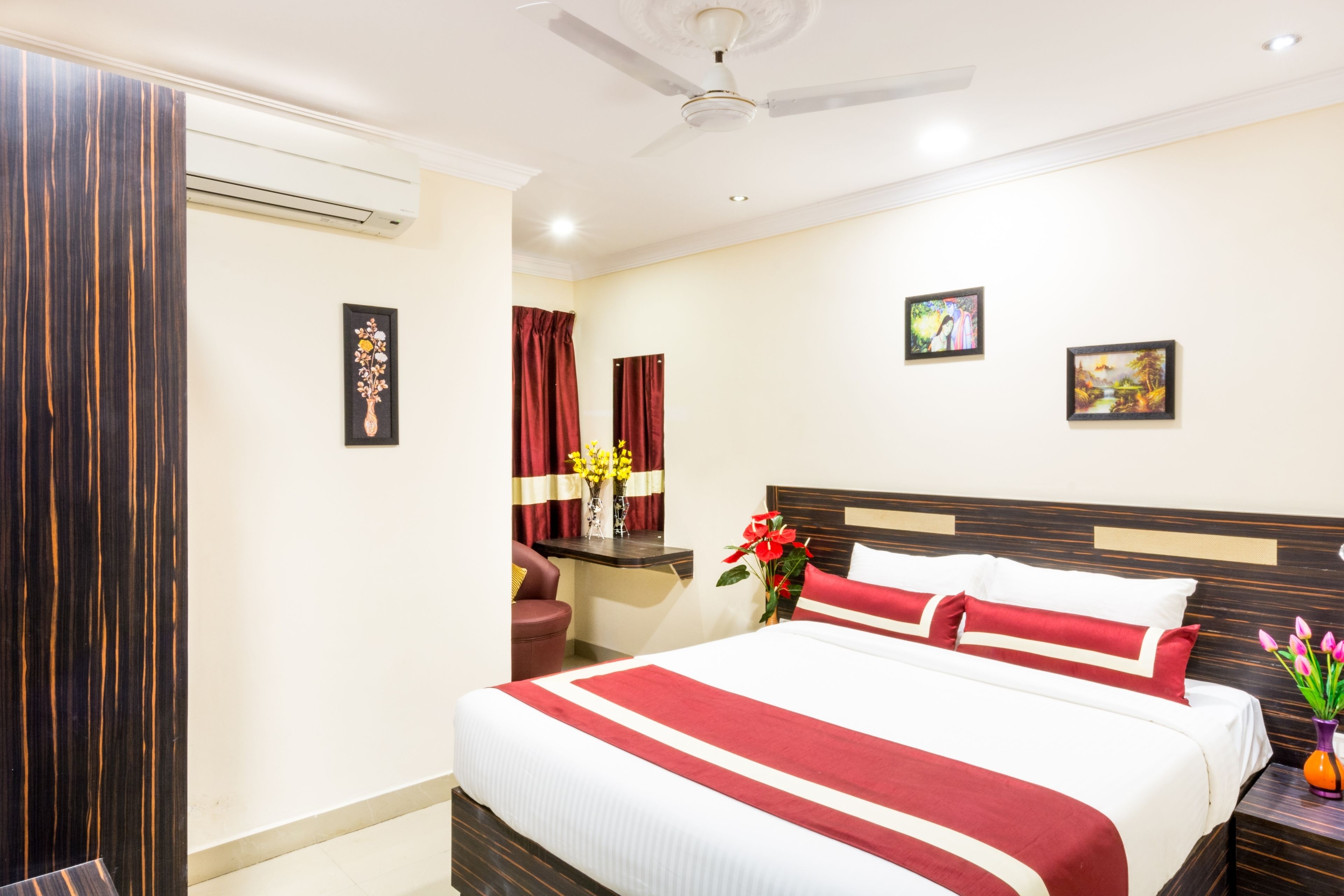 A hotel room with a large bed, a ceiling fan, a desk with a chair, and two paintings on the wall.