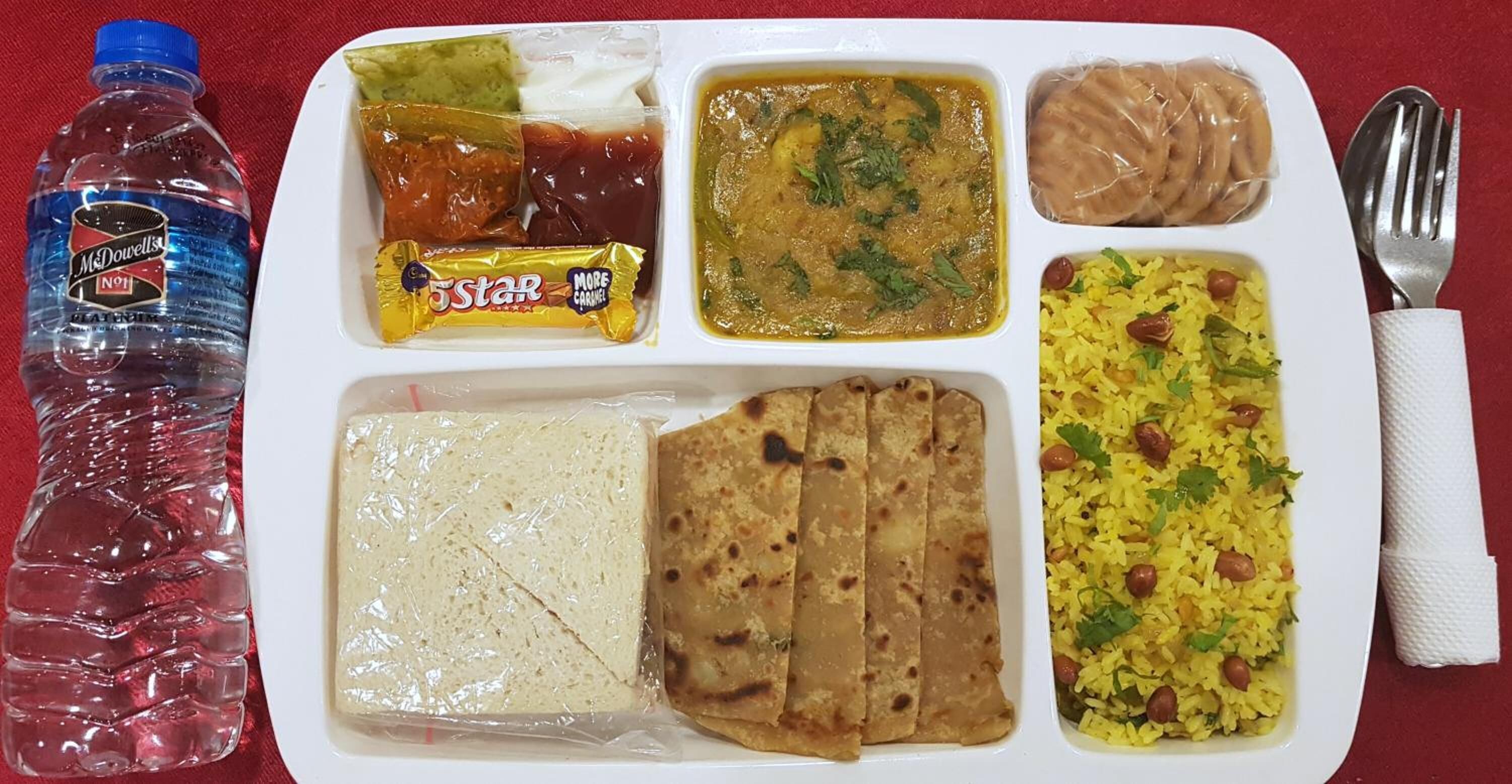 A meal tray with various dishes including rice, flatbread, and a curry, along with a bottle of water and a snack.