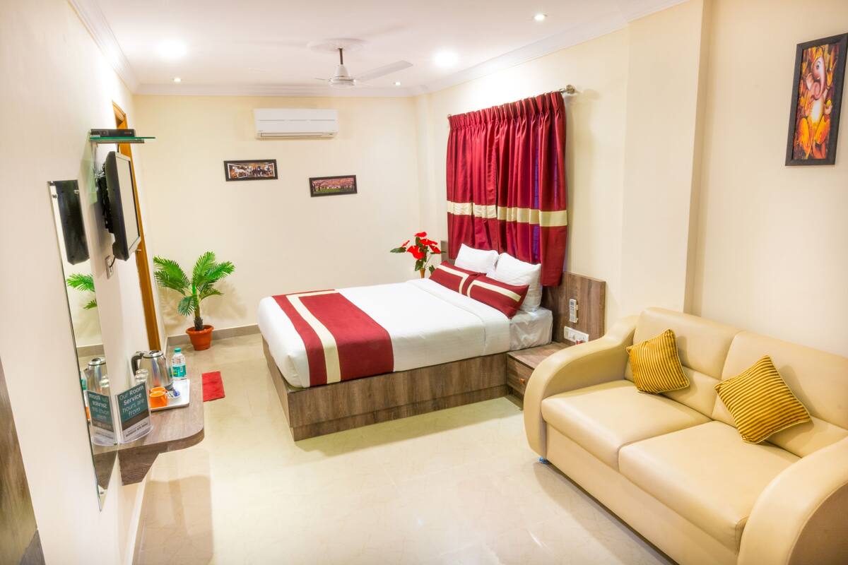 A hotel room with a bed, a sofa, a TV, a mirror, a plant, and a red curtain.