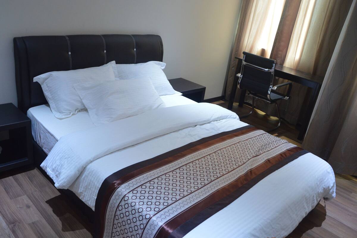Uma cama bem arrumada com lençóis brancos, cabeceira escura e manta decorativa estampada.