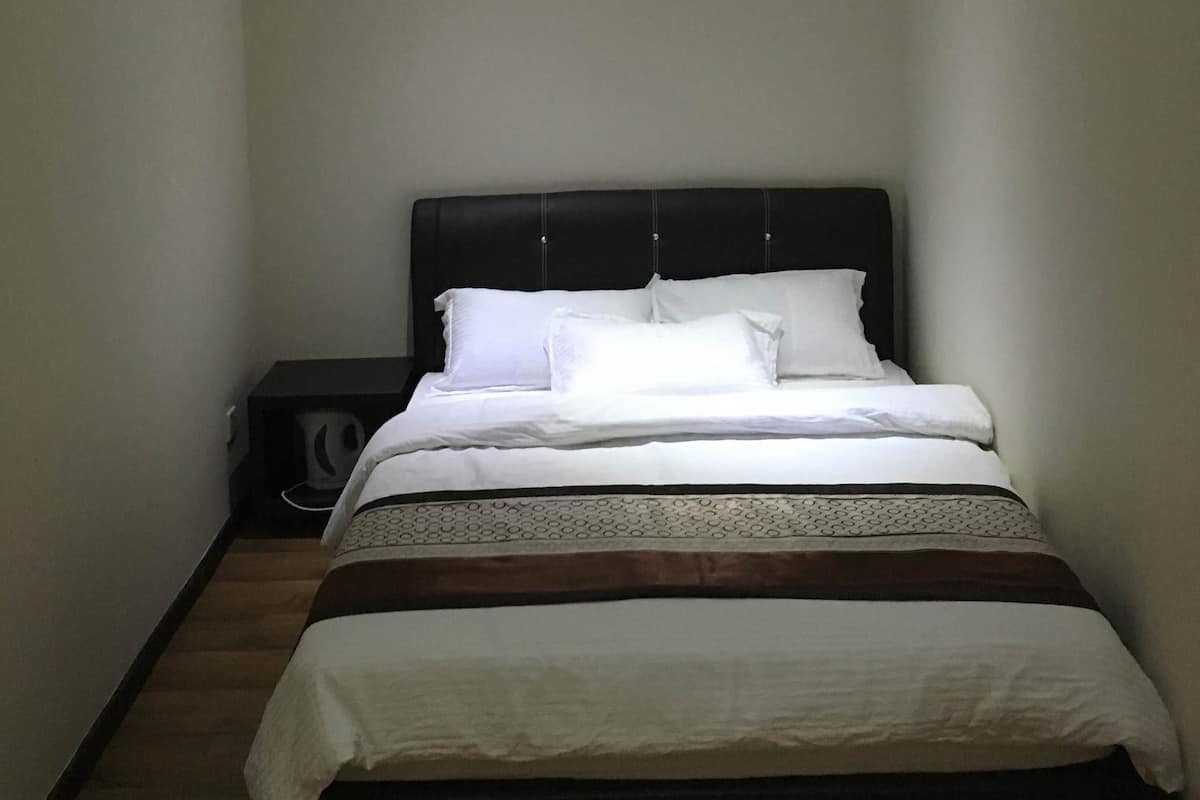 Quarto de hotel estreito com uma cama, uma mesinha de cabeceira e uma luminária fixada na parede.
