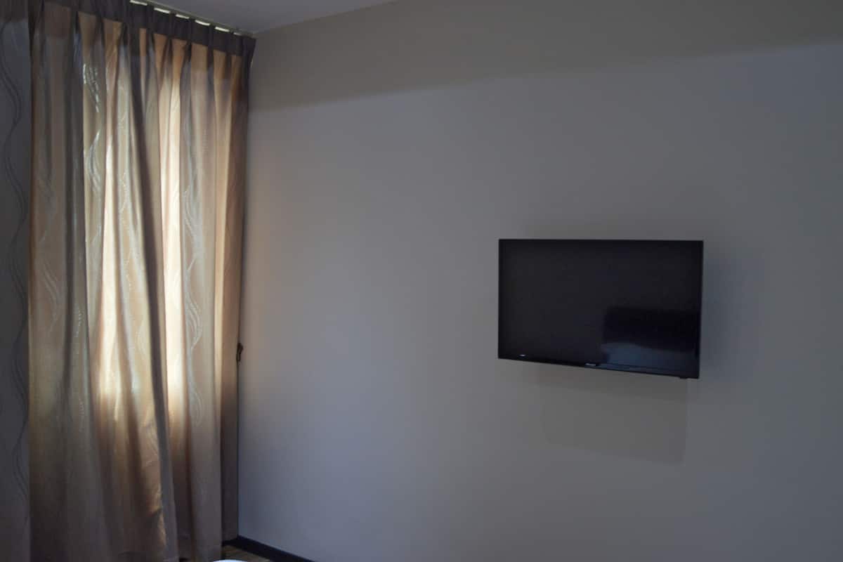 Quarto com televisão na parede, janela com cortinas e parede lisa.