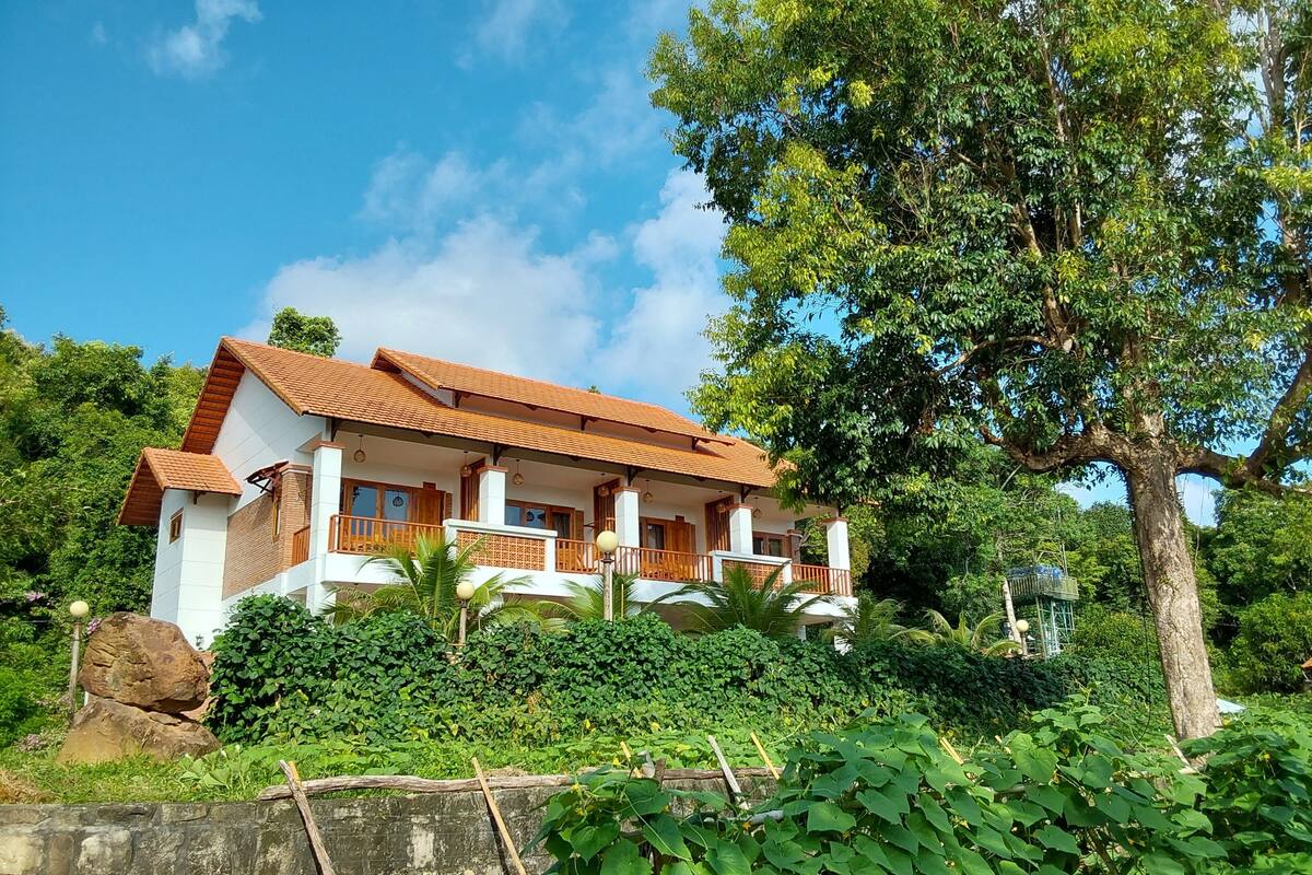 A two-story house with a tiled roof, surrounded by lush greenery and a large tree.
