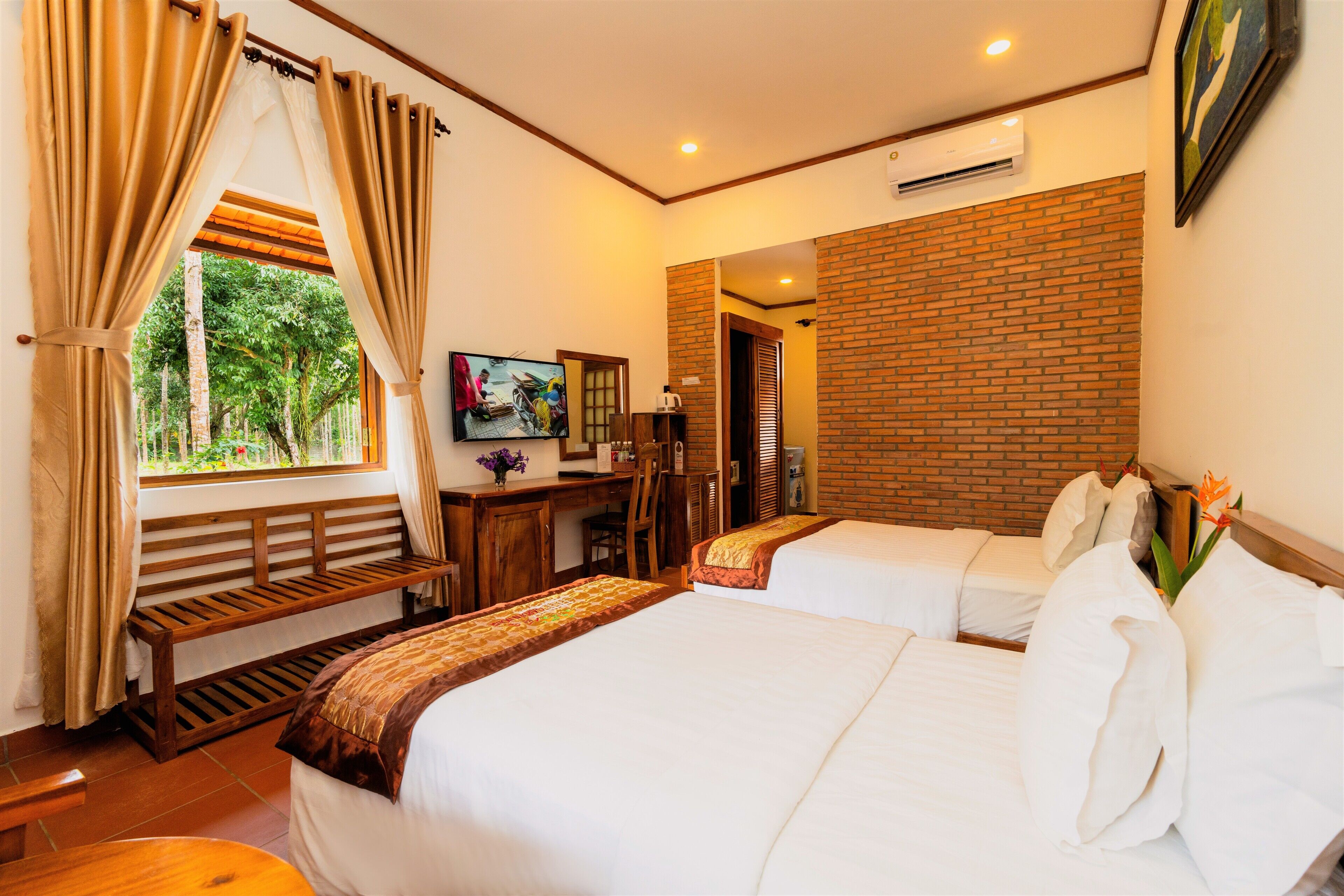 A hotel room with two beds, a desk, a chair, a window with curtains, a painting, and a brick wall.