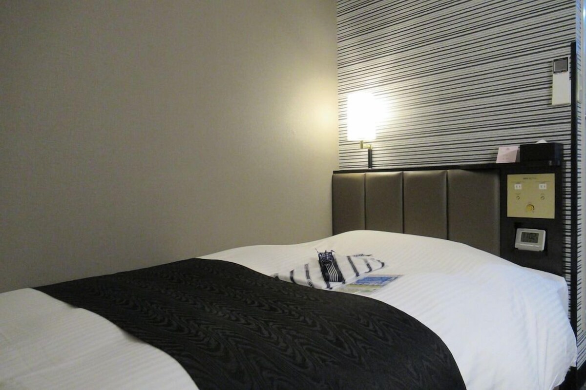 Habitación de hotel con cama, cabecera, panel de control en la pared y ventana con persianas.