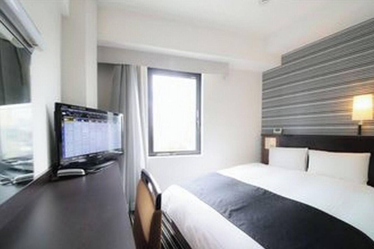 A hotel room with a bed, a desk with a computer, a window, and a wall-mounted light fixture.