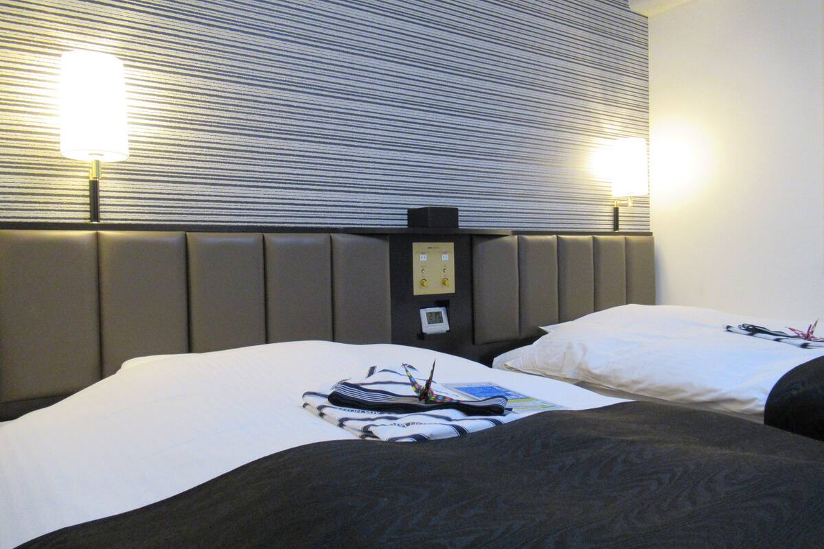 A hotel room with two beds, a headboard with a control panel, and wall-mounted lamps.