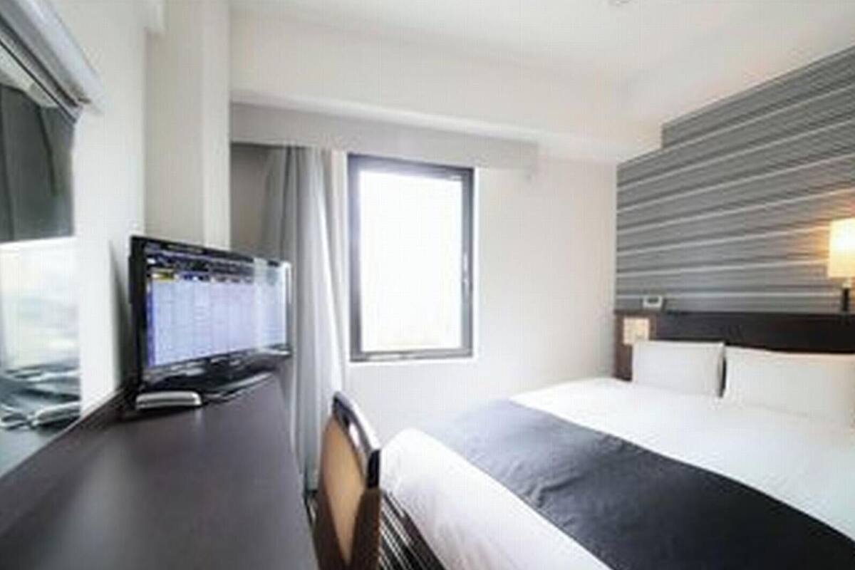 A hotel room with a bed, a desk with a computer, a window, and a wall-mounted light fixture.
