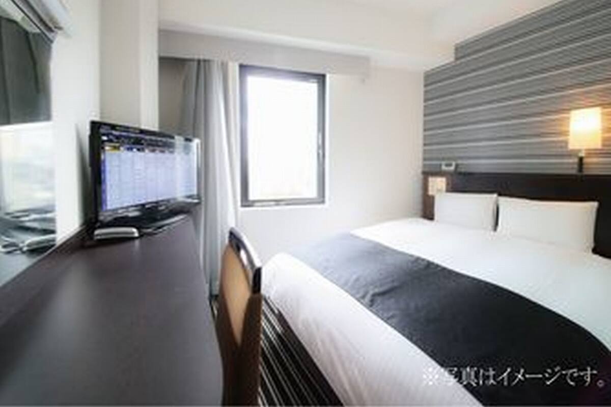 A hotel room with a bed, a desk with a computer, a window, and a wall-mounted light.