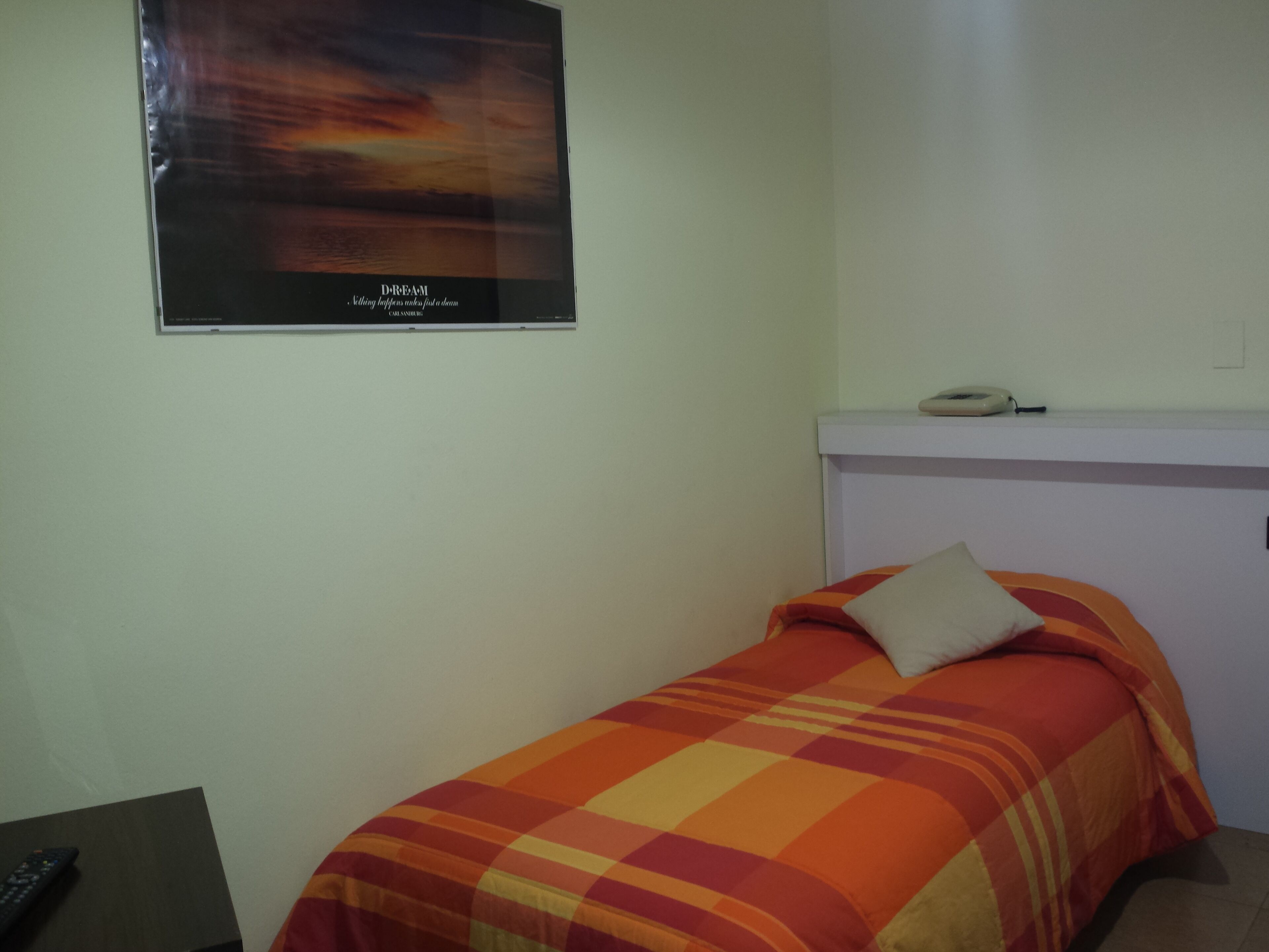 A single bed with a colorful blanket, a wall-mounted telephone, and a poster with text.