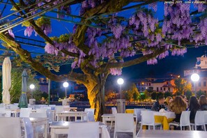 An outdoor dining area with tables and chairs, a large tree with purple flowers, and a cityscape in the background. - Hotel Stazione sul lago di Iseo (Paratico)