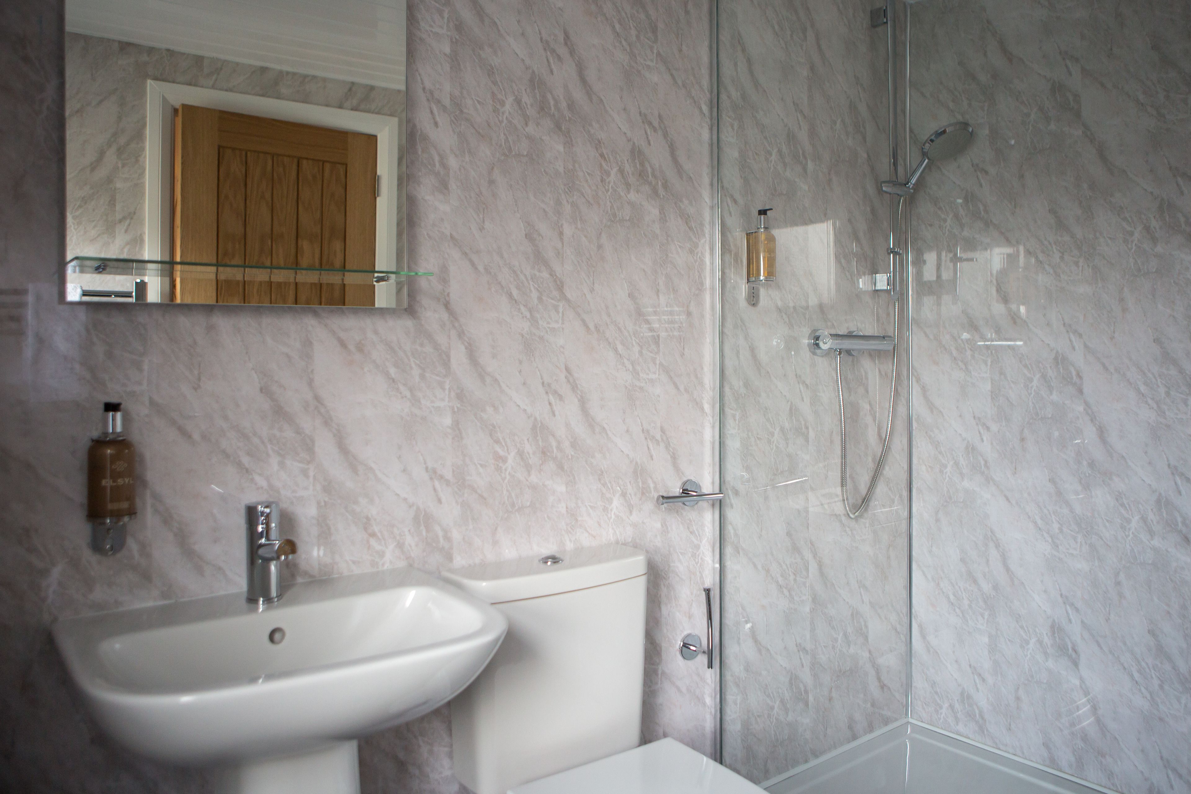 Un baño con pared de mármol, cabina de ducha de vidrio, lavabo blanco y puerta de madera.