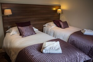 A hotel room with two beds, each with a headboard, pillows, and a folded towel on the bed. There are wall-mounted lamps on either side of the headboard. - Foundry 34 (Penrith)