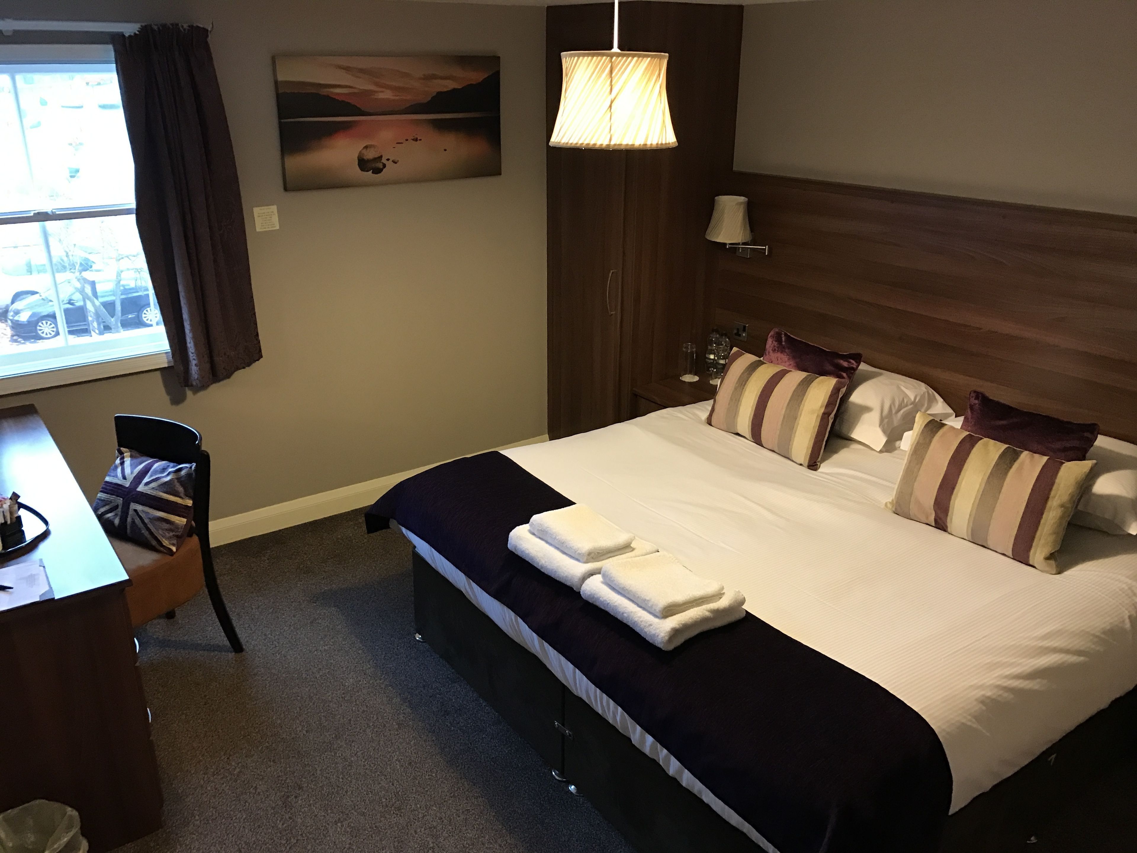 A hotel room with a large bed, a desk, a chair, a window with curtains, a painting on the wall, and a lamp.