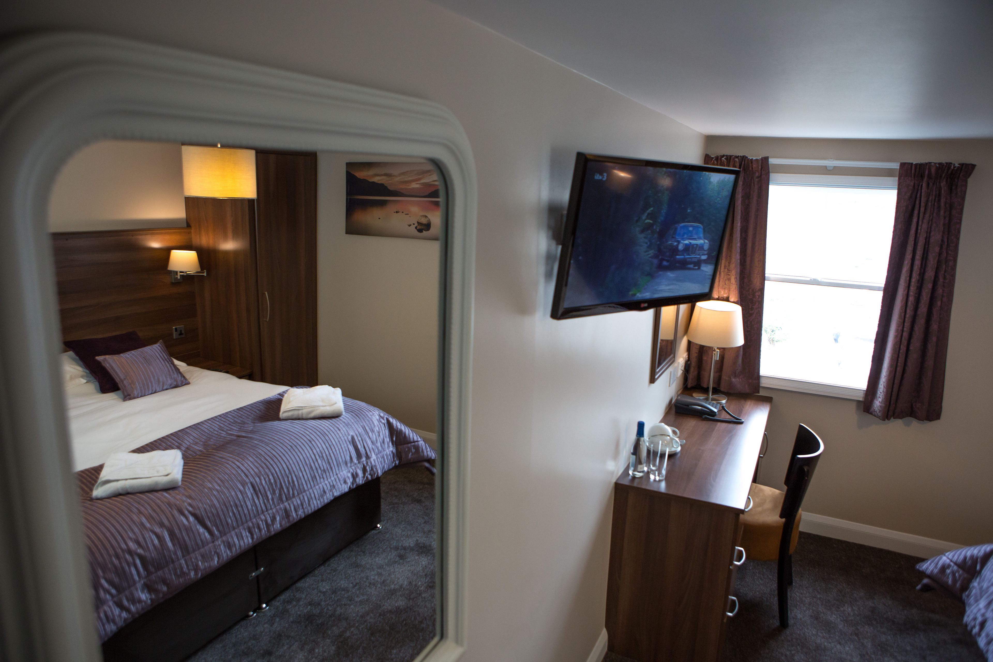 Quarto de hotel com cama, escrivaninha, cadeira, televisão e janela com cortinas.