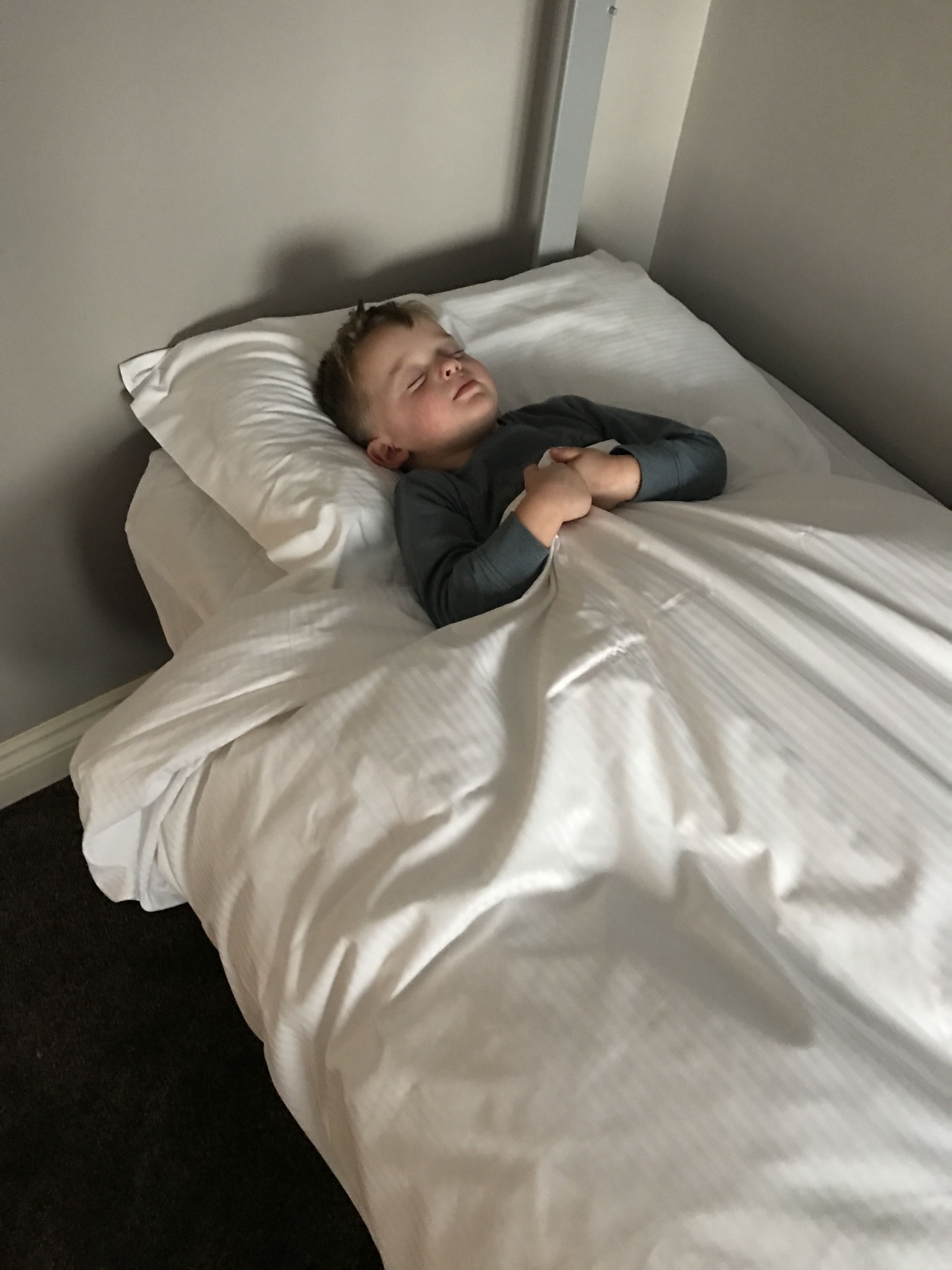A child lying in a bed with white bedding in a room with a plain wall and a door.