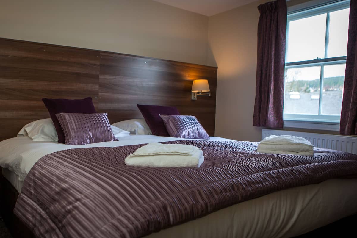 A hotel room with a large bed, two wall-mounted lamps, a window with curtains, and a view of boats outside.