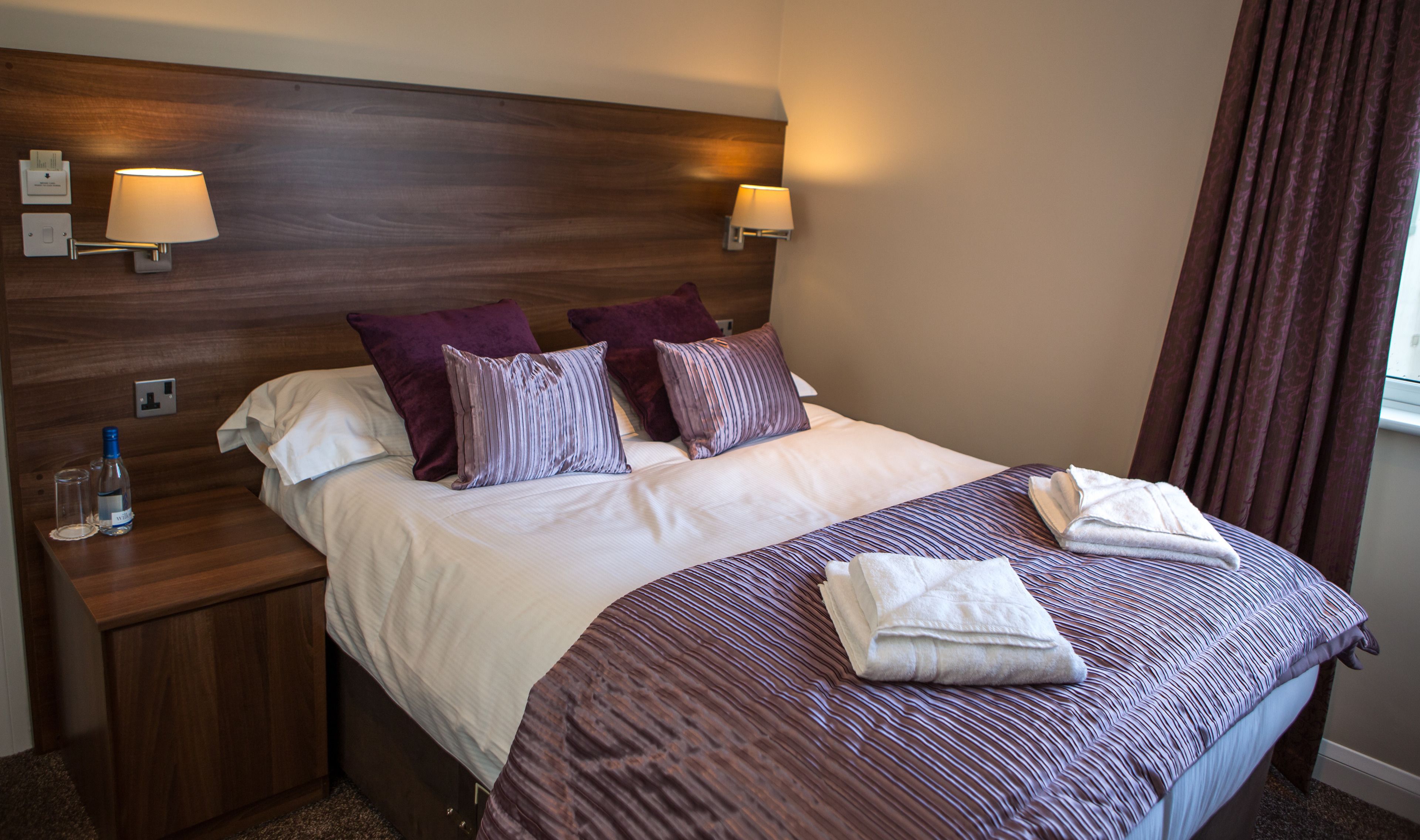 A hotel room with a wooden headboard, two bedside lamps, a bed with white and purple bedding, and a window with curtains.