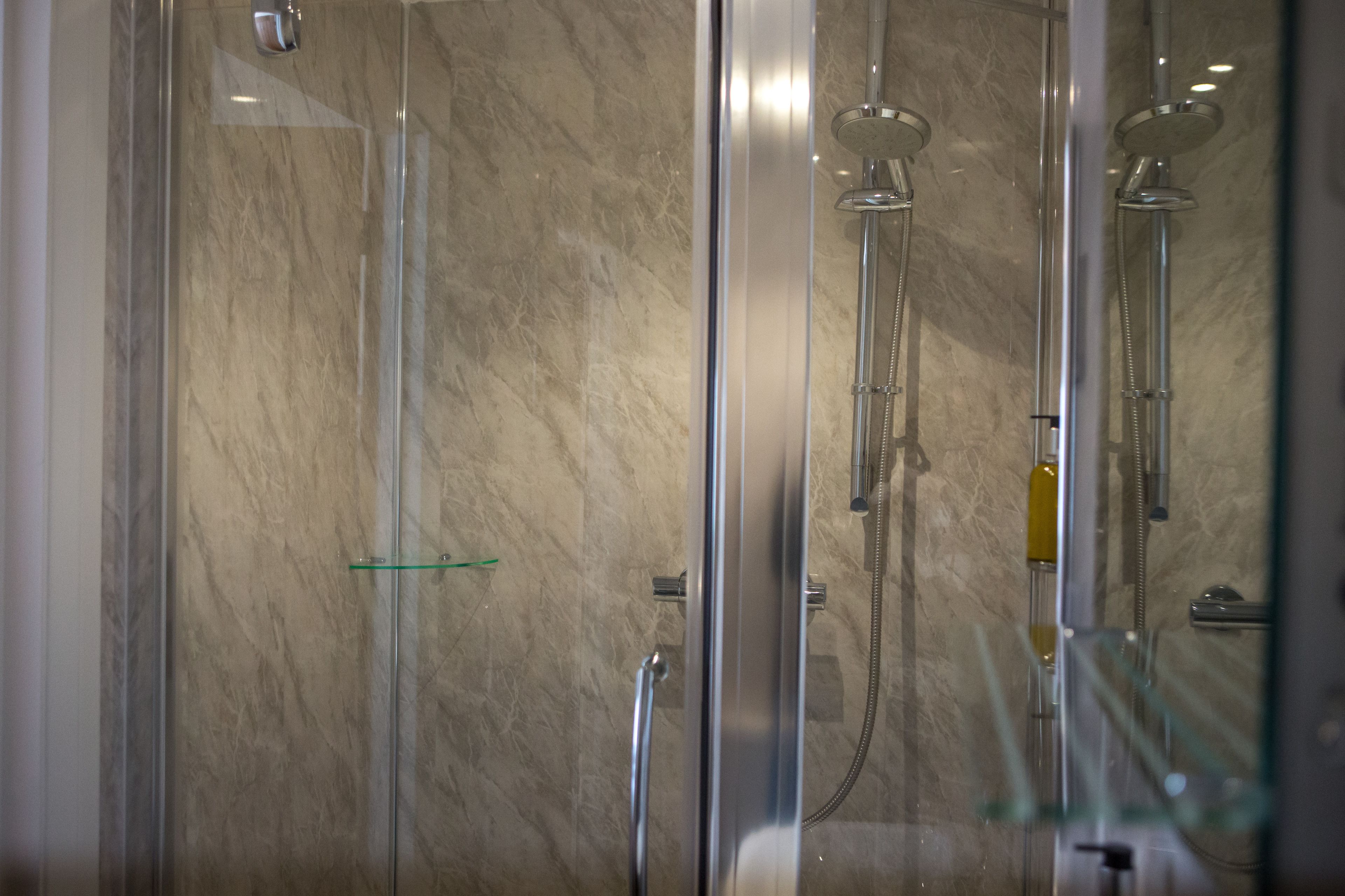 A glass-enclosed shower with a marble wall, a glass shelf, and a handheld showerhead.