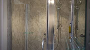 A glass-enclosed shower with a marble wall, a glass shelf, and a handheld showerhead.