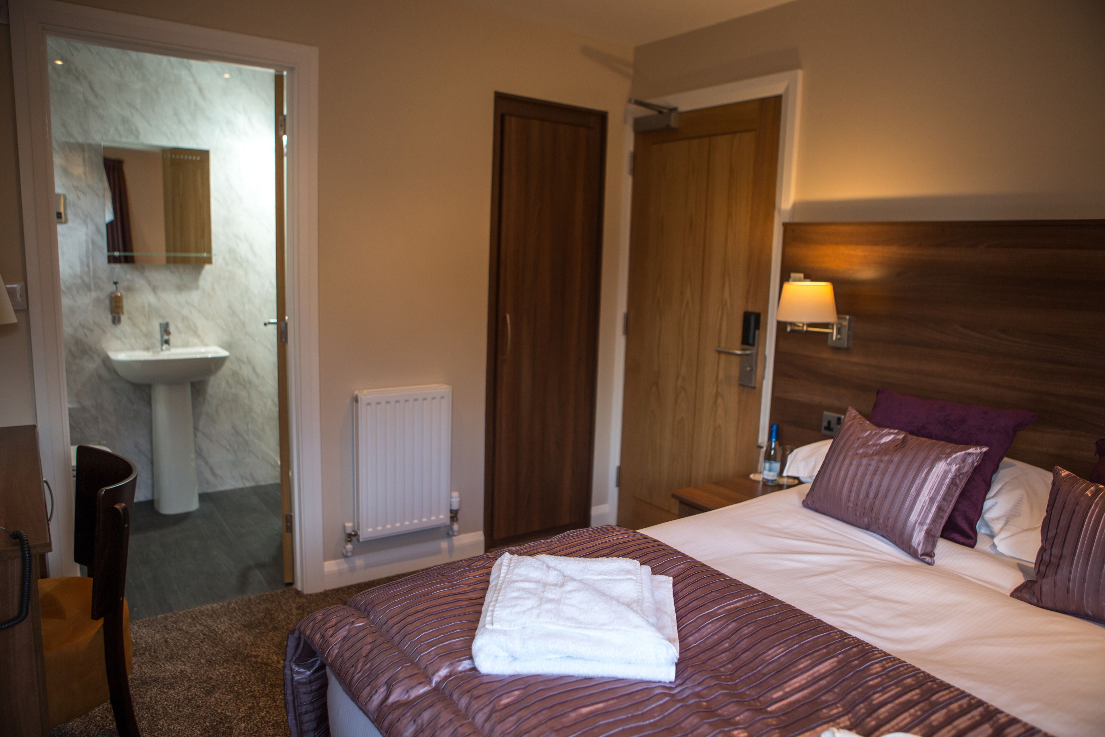 Ein Hotelzimmer mit Bett, Holz-Kopfteil, Nachttisch mit Lampe, Badezimmer mit Waschbecken und Spiegel sowie einer Tür zu einem anderen Raum.