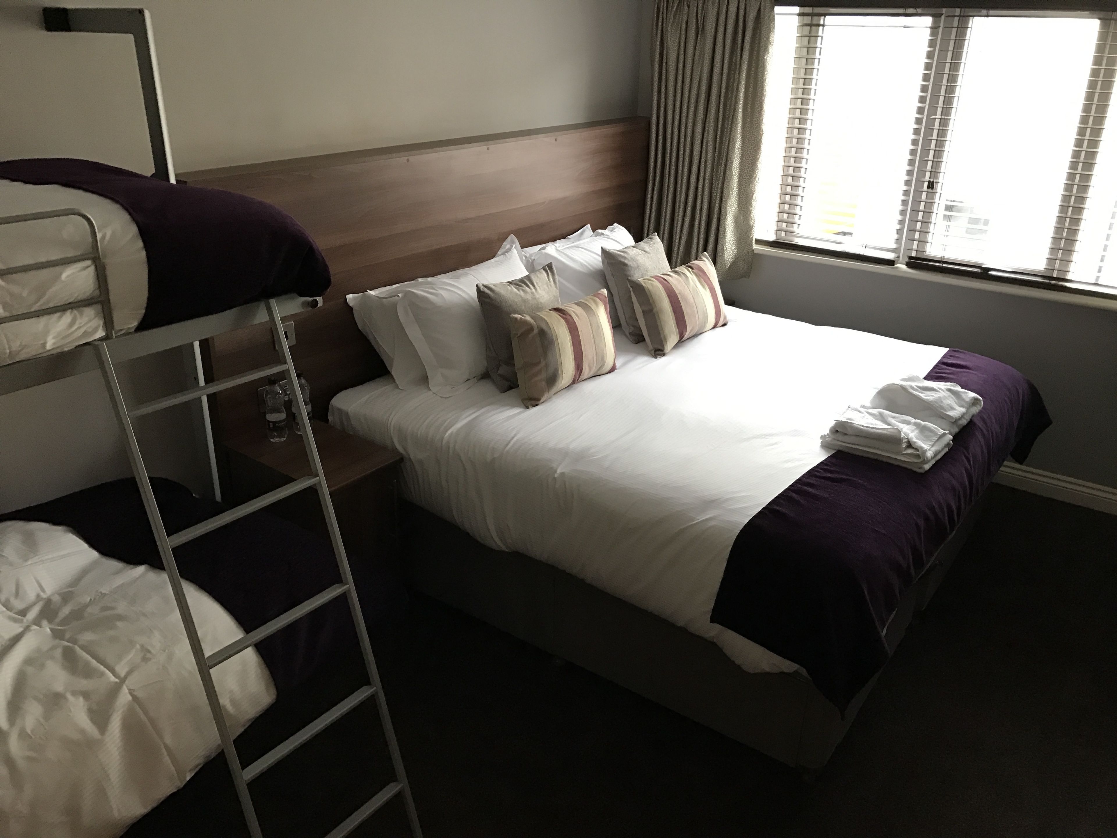 A hotel room with a bunk bed, a single bed with a wooden headboard, and a window with curtains.