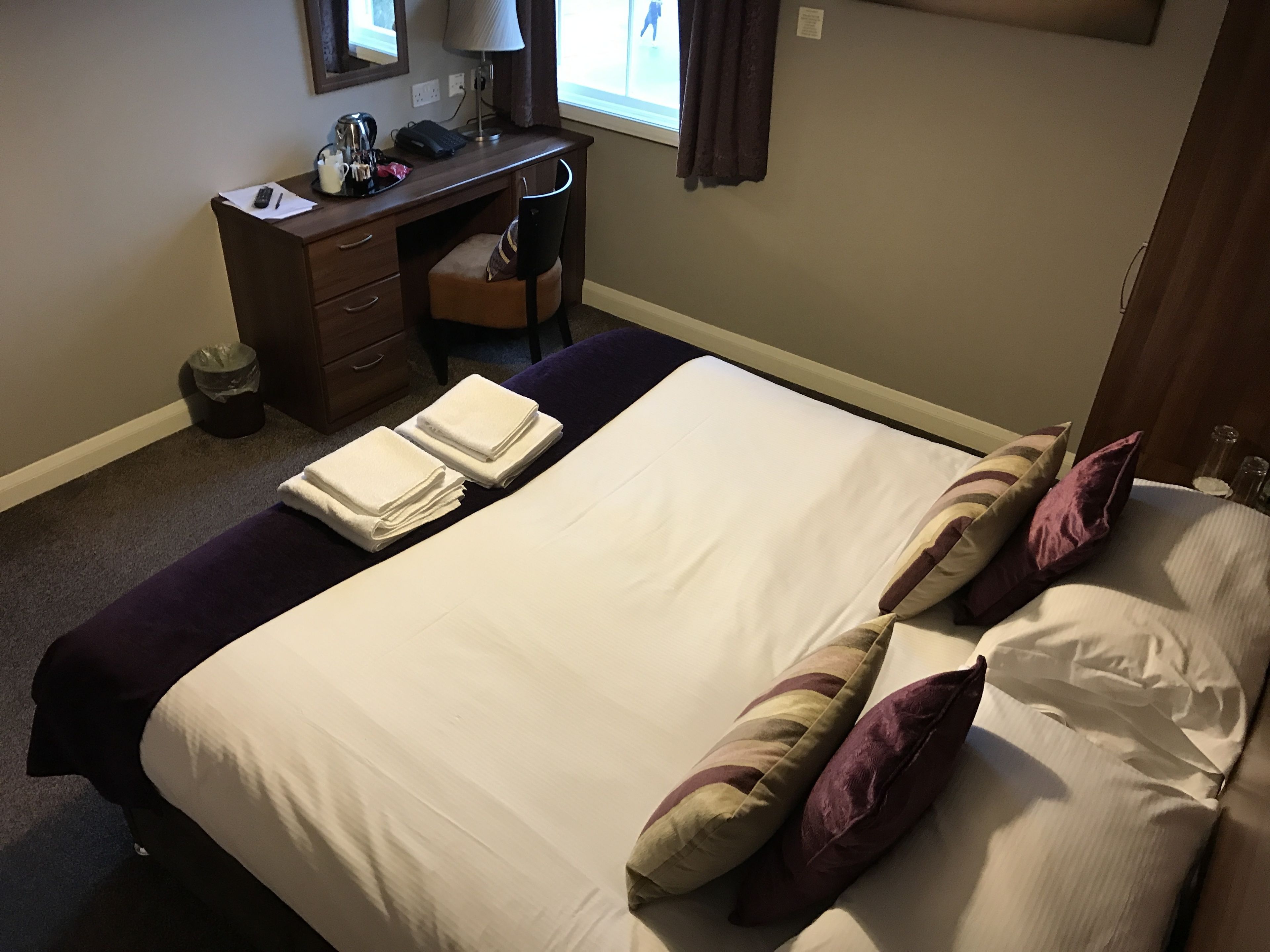 A neatly made bed with pillows, a desk with a lamp, and a chair.