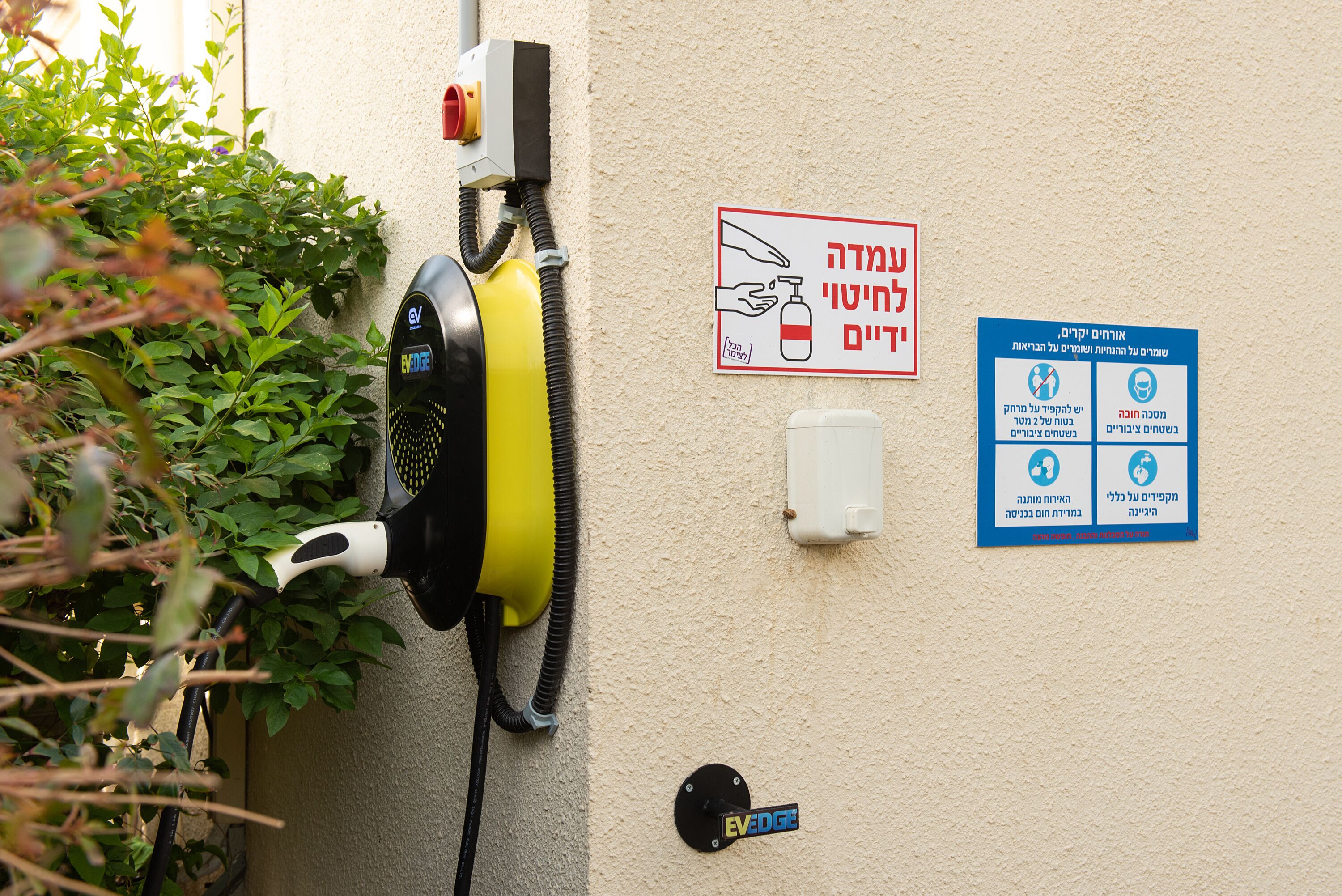 Une borne de recharge publique fixée au mur, de couleur jaune et noire, équipée d’un distributeur de gel hydroalcoolique et de panneaux d’information en hébreu.