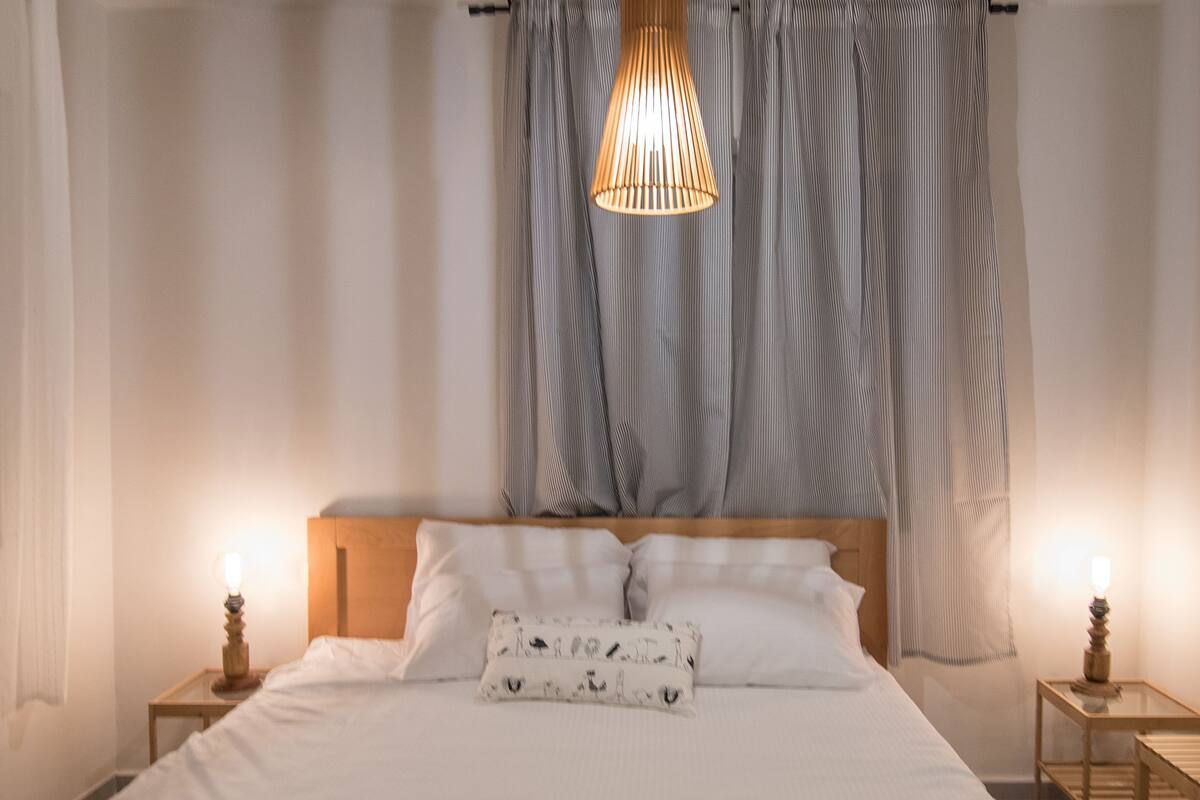 A neatly made bed with white linens, a wooden headboard, and a hanging light fixture. There are two bedside tables with lamps.