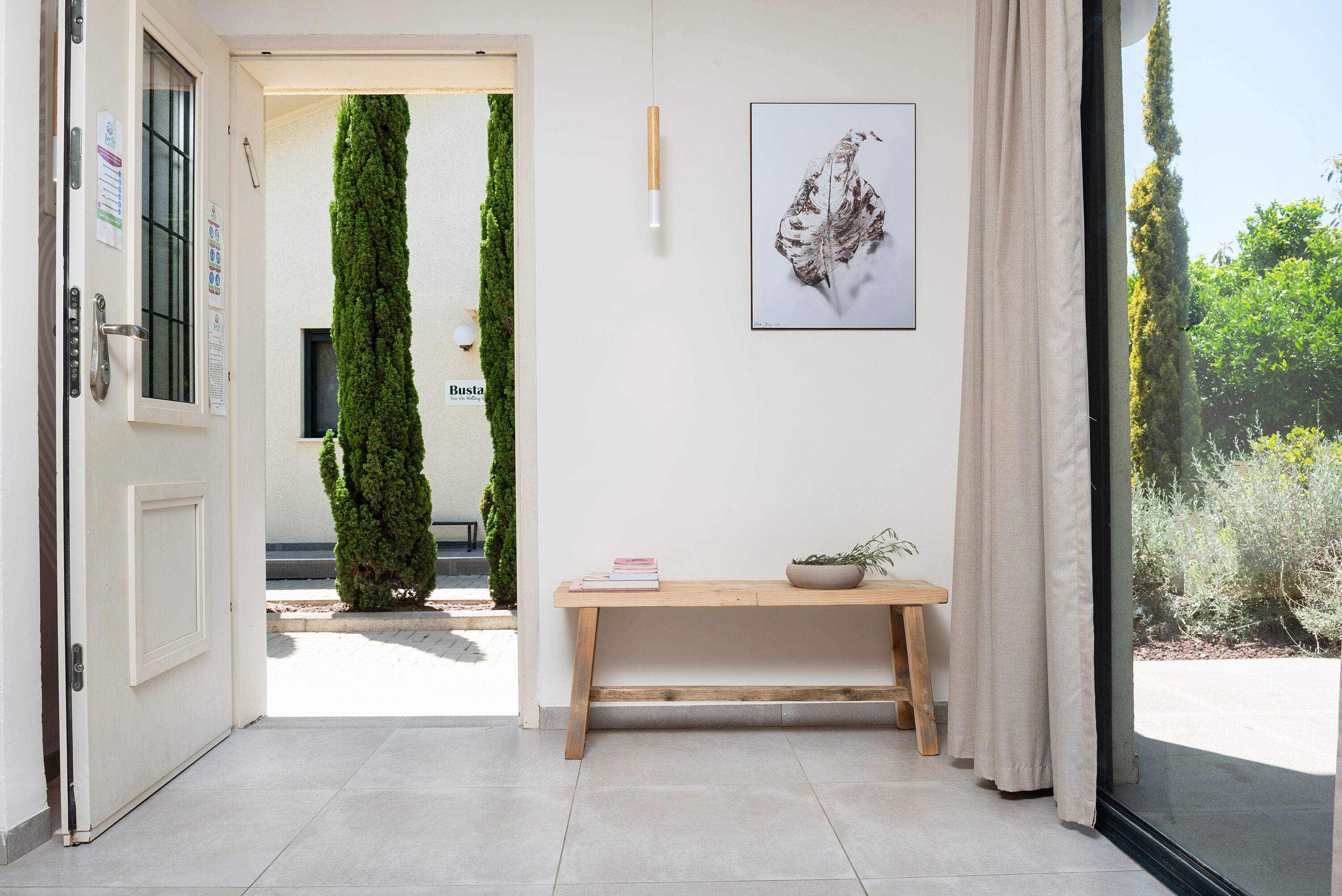 A modern interior with a wooden bench, a framed artwork, and a door leading to an outdoor area with a green wall and a small tree.