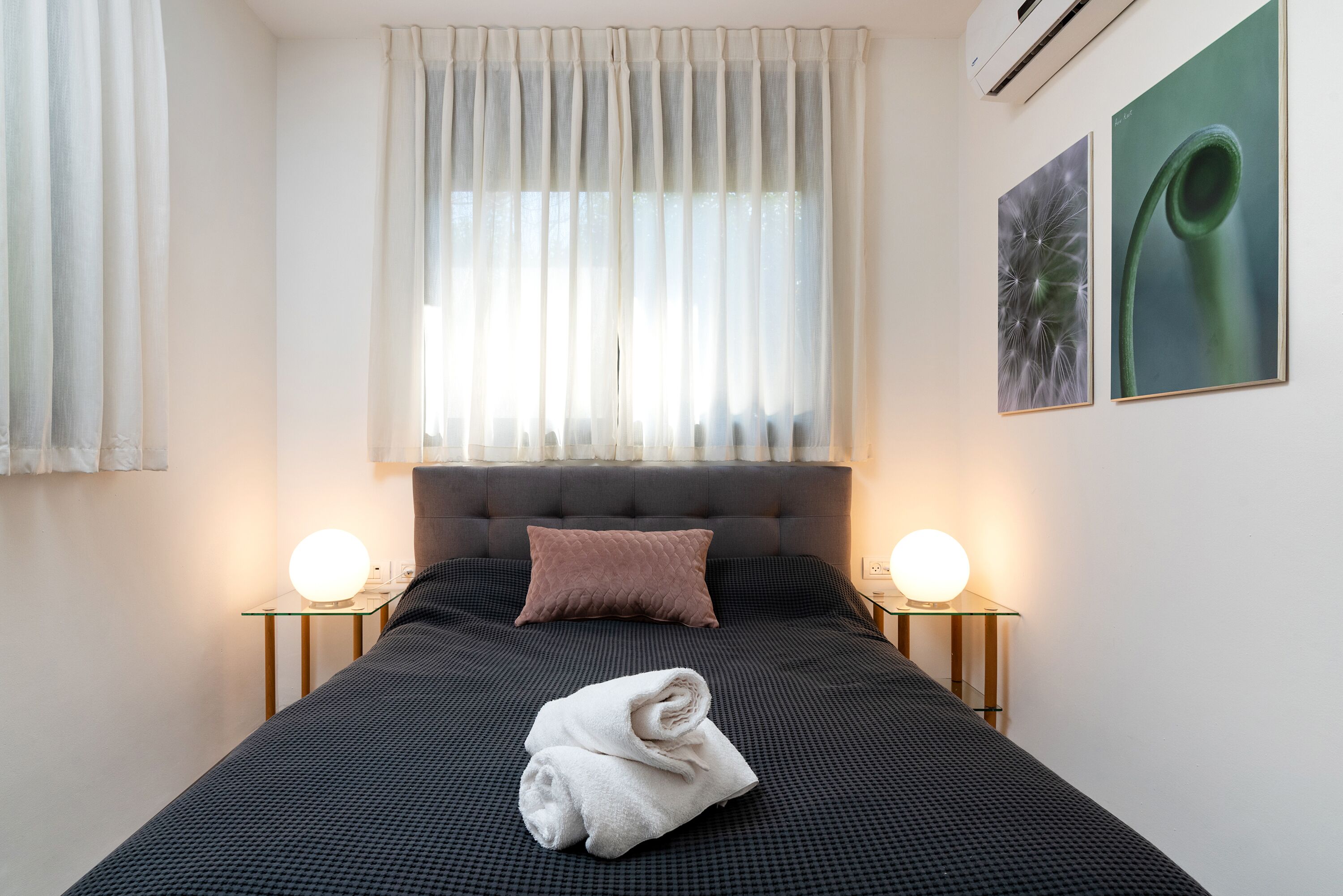 A neatly made bed with a folded towel, two bedside lamps, a window with sheer curtains, and two framed pictures on the wall.
