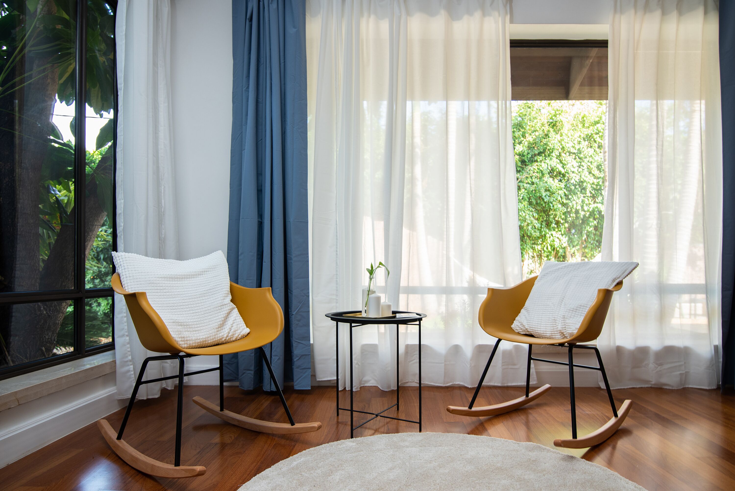 Una habitación con dos mecedoras, una mesita con un florero y vistas a la vegetación a través de cortinas traslúcidas.