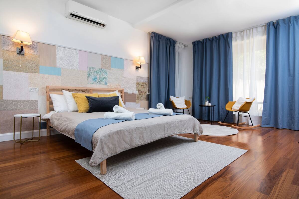 A modern hotel room with a large bed, a small side table, a rocking chair, and a large window with curtains.