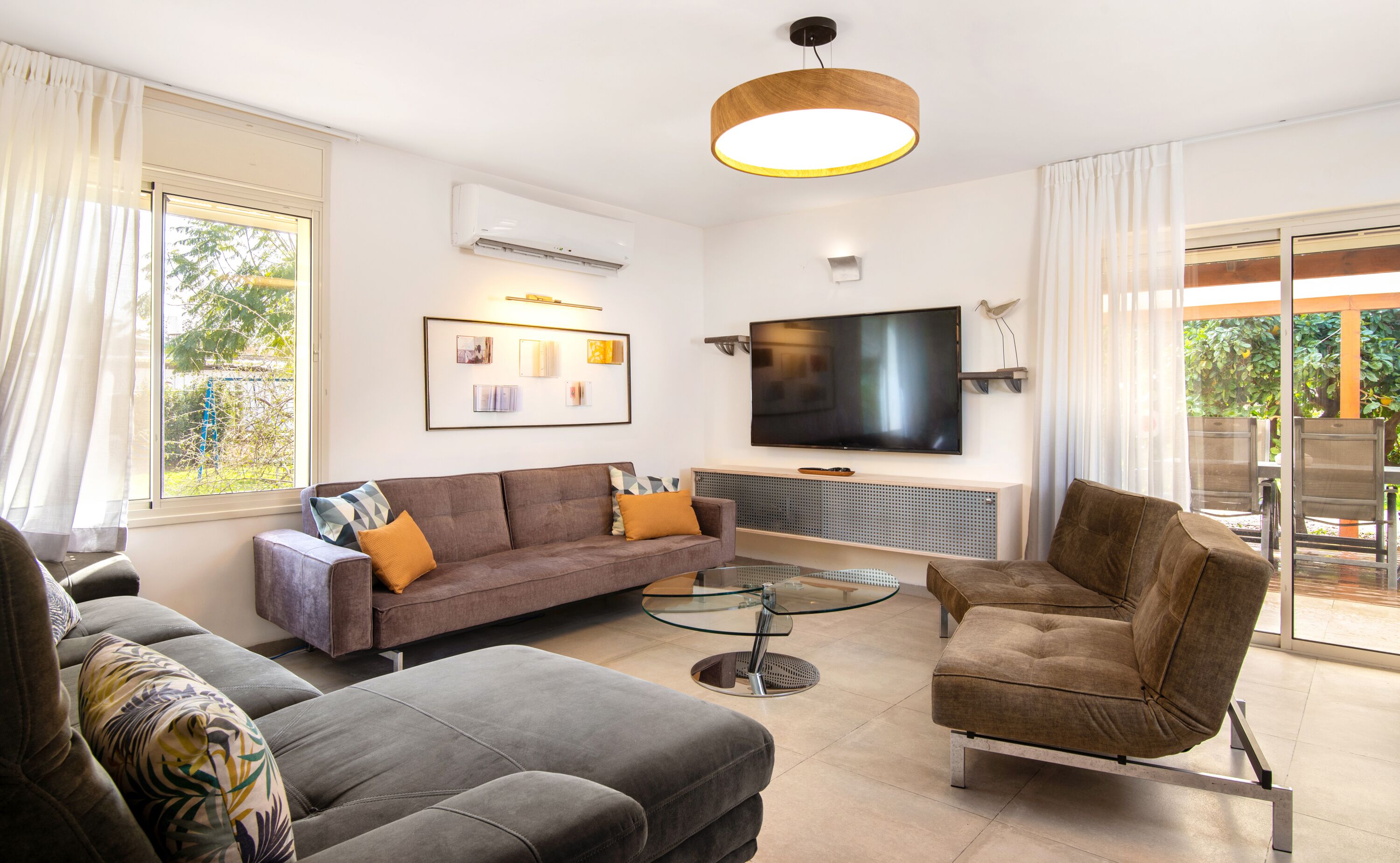 A modern living room with a sofa, armchair, and glass coffee table. A flat-screen TV is mounted on the wall, and there is a ceiling light fixture.