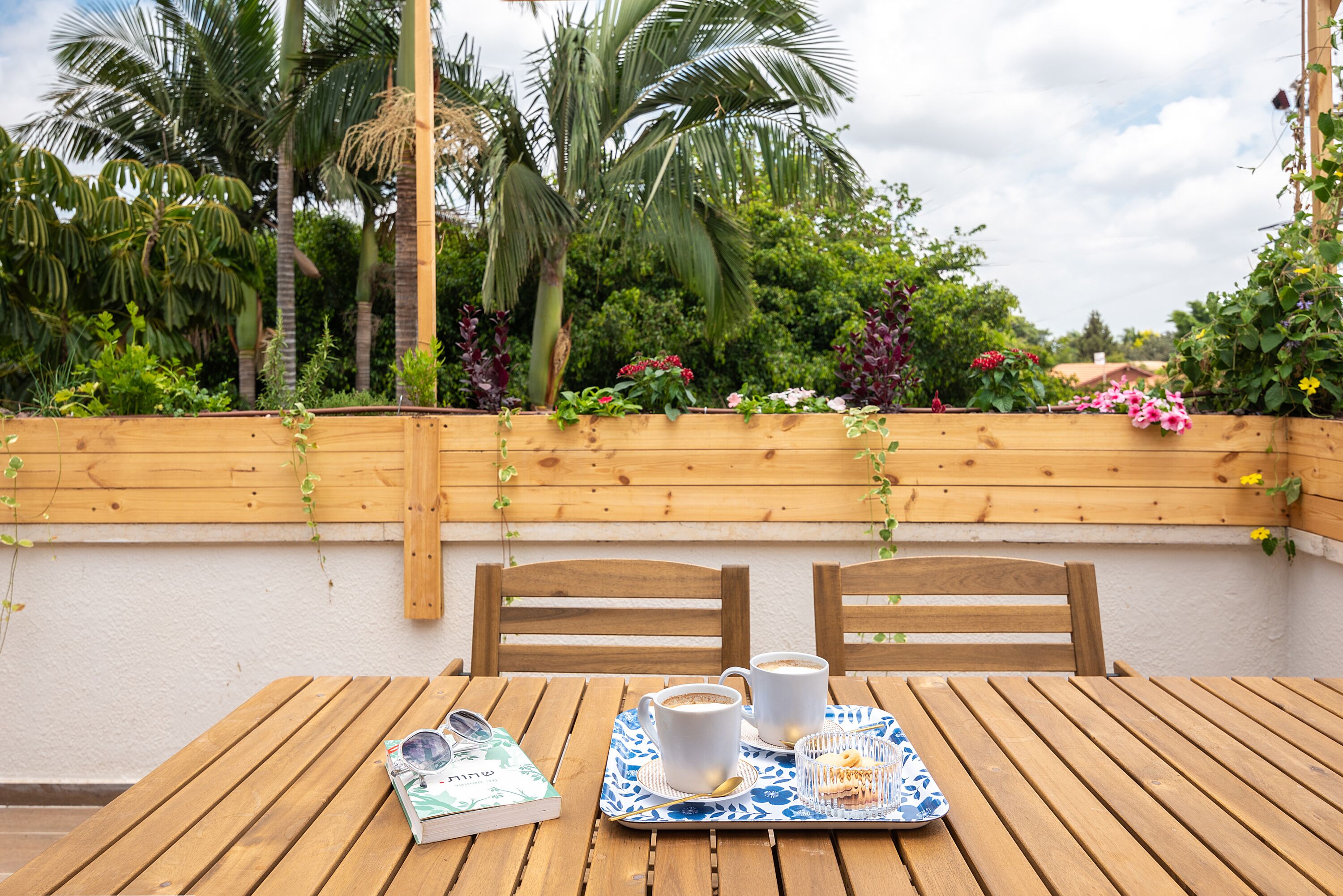 Uma mesa de madeira com duas cadeiras, um tabuleiro com uma xícara e um livro, e uma cerca de madeira com plantas e flores.