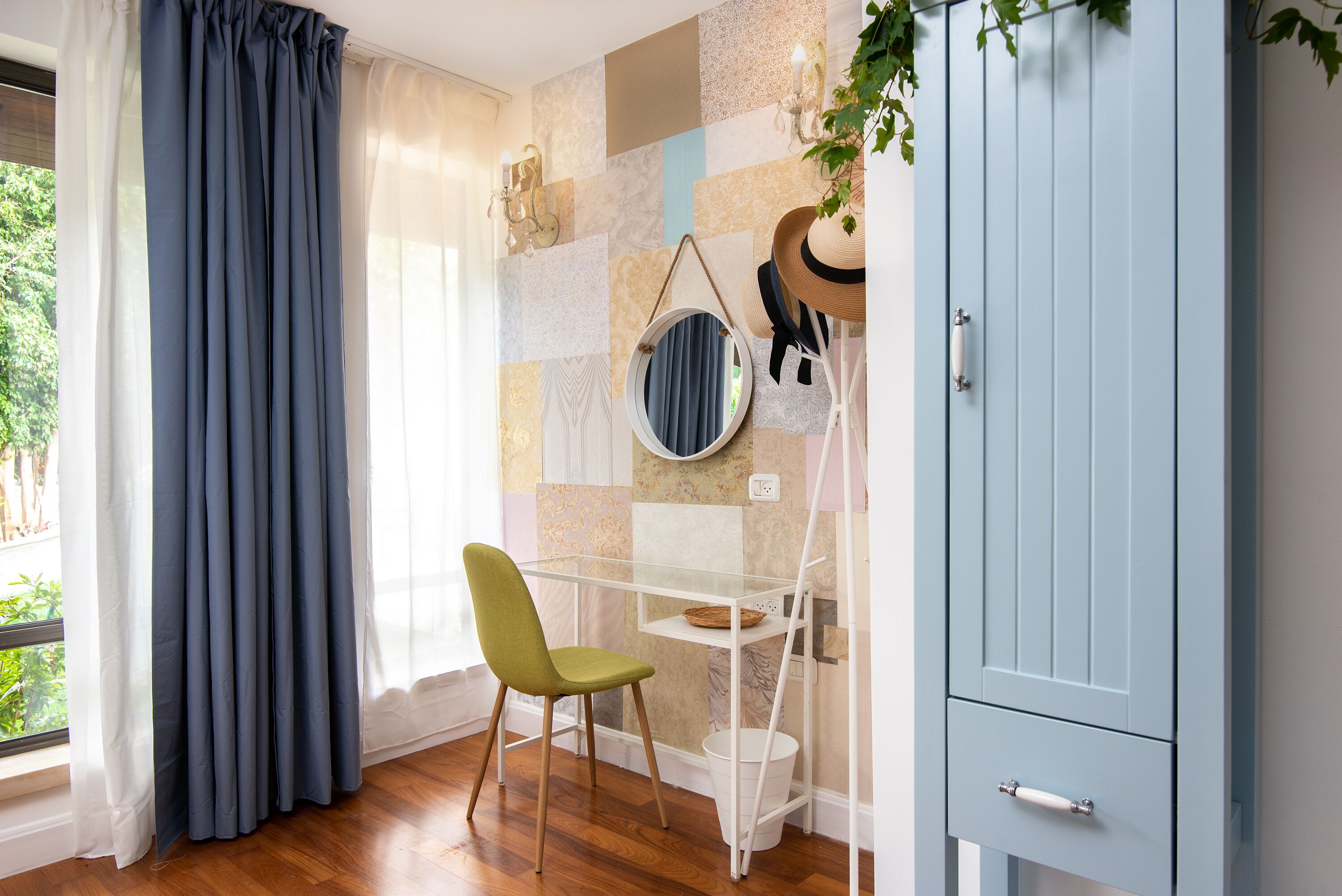 Een kamer met houten vloer, een bureau met stoel, een kledingkast, een raam met gordijnen, en een wand met een decoratief patroon.