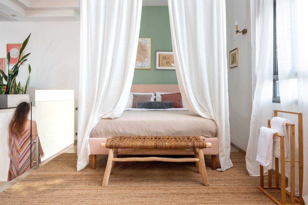 A bedroom with a bed, a bench, a bedside table, a plant, and a window with curtains.