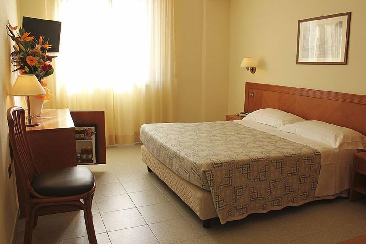 Habitación de hotel con cama, escritorio con lámpara, silla, televisor y un cuadro en la pared.