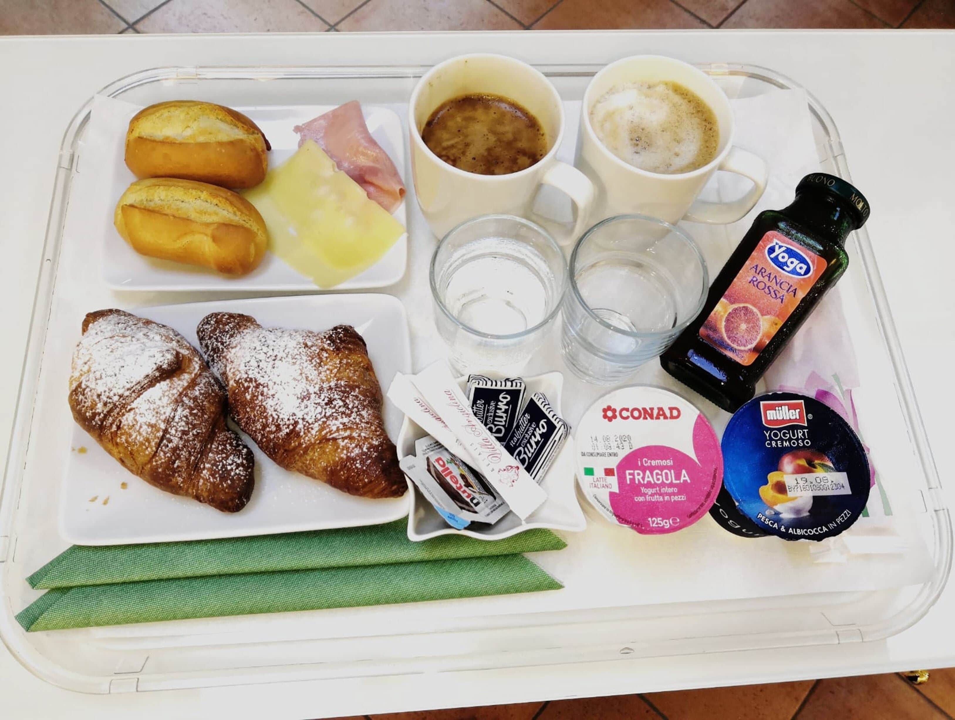 Ein Tablett mit Croissants, Käse, Schinken, Kaffee, Wasser, einer Flasche Yoga Arancia Rossa, CONAD Joghurt und einem Müller-Joghurtbecher.