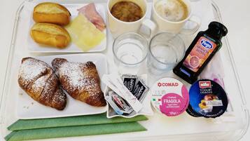 Ein Tablett mit Croissants, Käse, Schinken, Kaffee, Wasser, einer Flasche Yoga Arancia Rossa, CONAD Joghurt und einem Müller-Joghurtbecher.