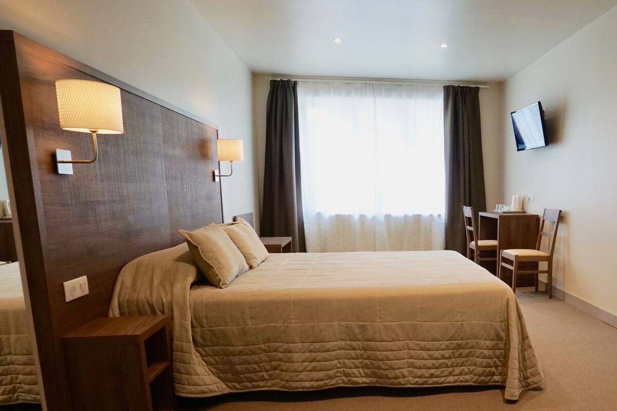 A hotel room with a large bed, a desk, a chair, a TV mounted on the wall, and a window with curtains.