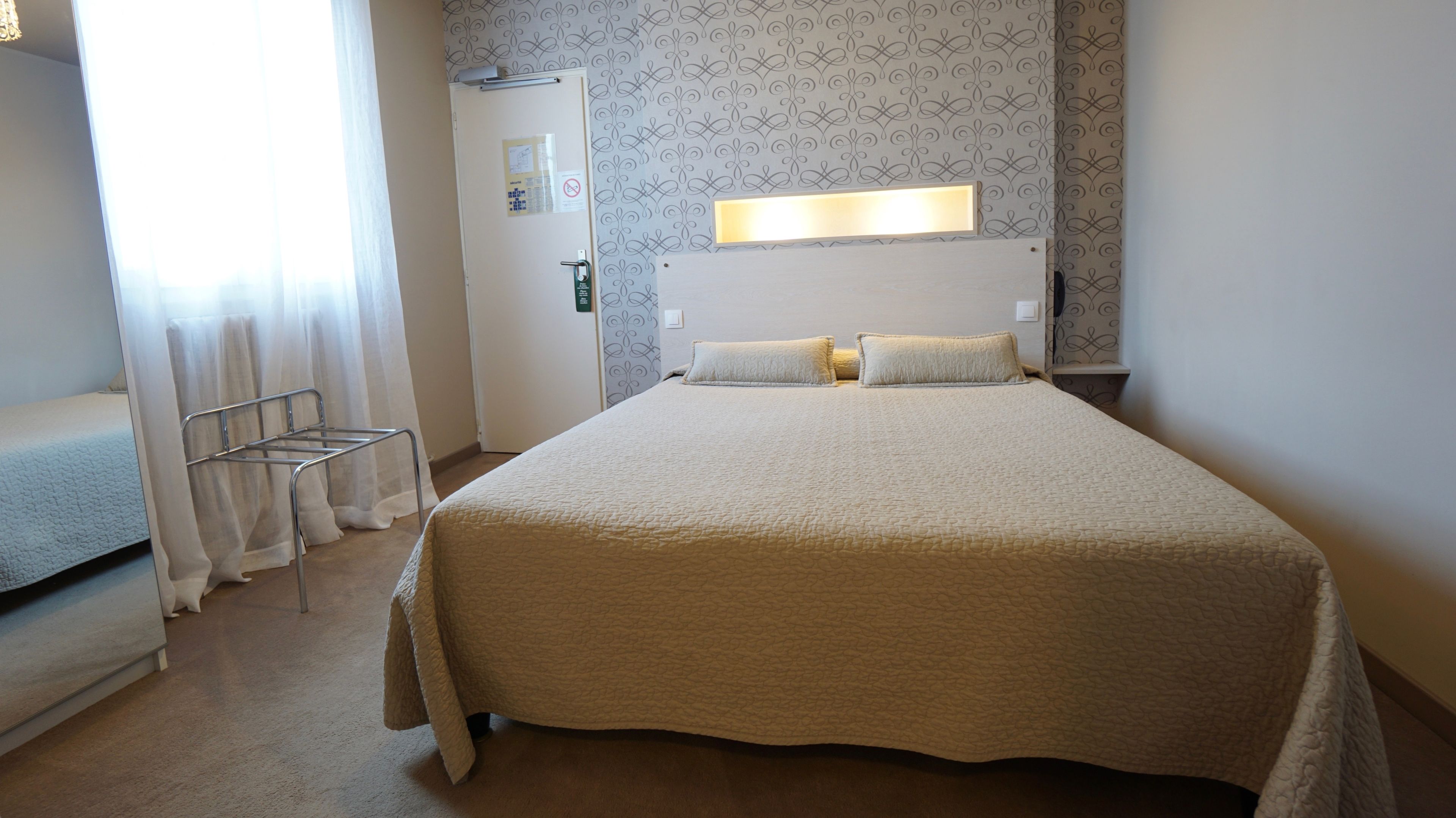 A hotel room with a large bed, a bedside table, a chair, and a door with a light switch.