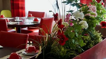 Un comedor con sillas y mesas rojas, una planta en maceta con flores y una cortina roja al fondo.